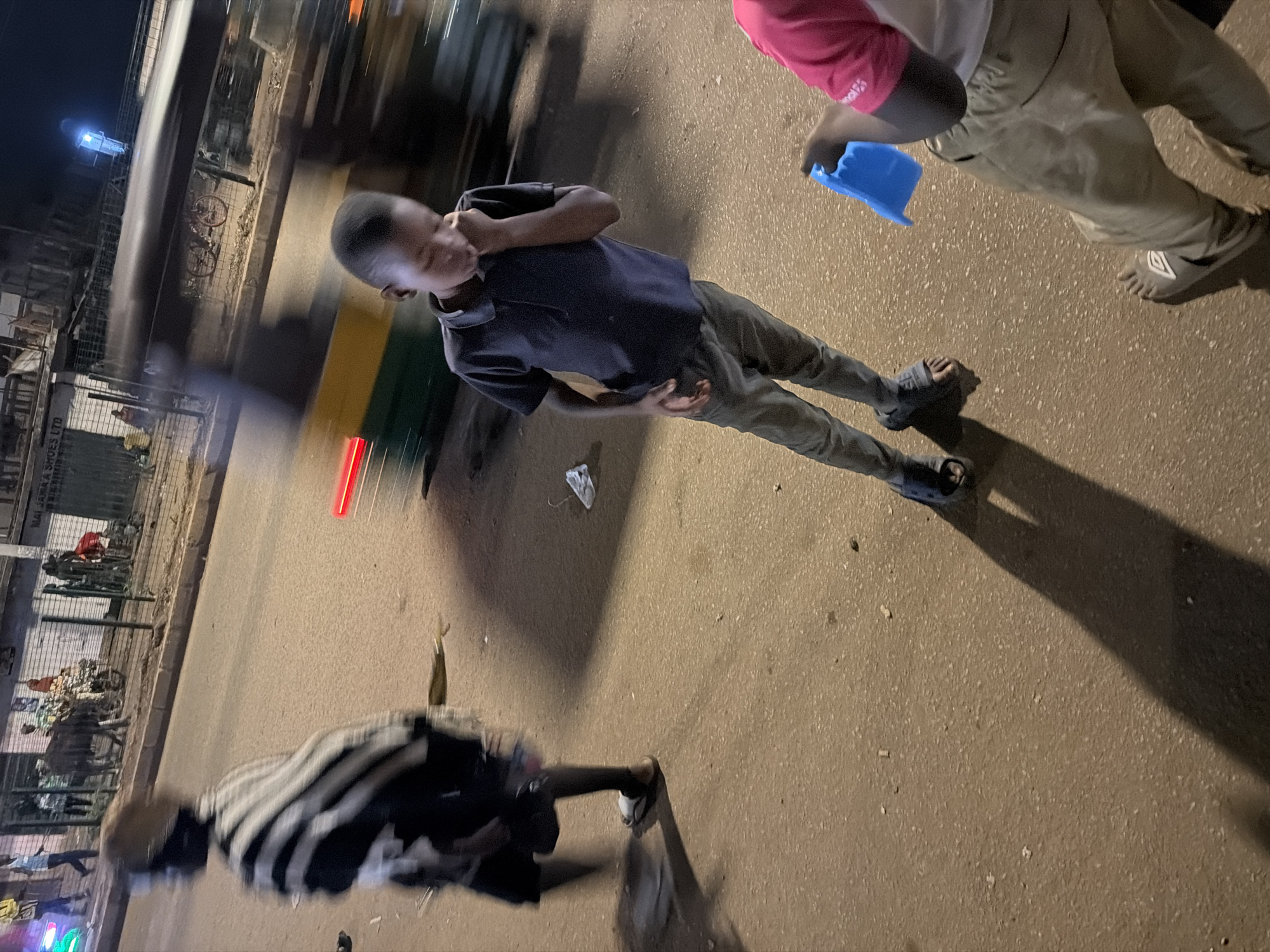 The image depicts a street scene in Bukuru, Nigeria at night. In the foreground, a young boy in a dark polo shirt and grey pants stands with his hand to his mouth, appearing to be thinking or possibly eating. He is wearing sandals. To his right, another child, partially visible, wears a pink shirt and tan pants and holds a blue plastic object. Further left and blurred due to motion, a person in a black and white striped garment walks away from the camera. The background shows a street with a moving vehicle that is blurred, indicating it is in motion. Buildings line the street, one with signage that includes "MAIJAMA'A SHOES LTD." The street is paved with asphalt, and there are some scattered debris. The lighting suggests nighttime, with artificial lights illuminating the scene and casting long shadows. There are also animals, possibly cows, and motorcycles visible in the distance, contributing to the impression of a bustling urban environment.