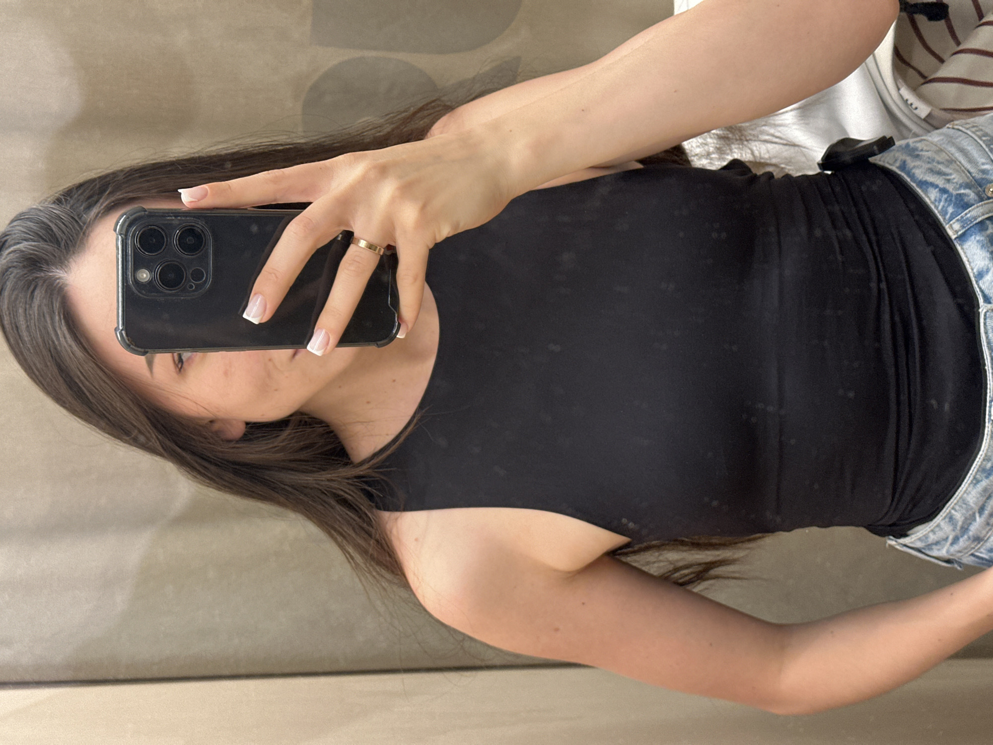 A woman stands in a changing room, holding a black smartphone up to take a selfie. She is wearing a black tank top and light-wash denim shorts. Her hair is brown with some gray at the roots. The changing room has neutral-toned walls. The image appears to be taken indoors. There is no verifiable evidence that the photo was taken in Kazan, Russia.