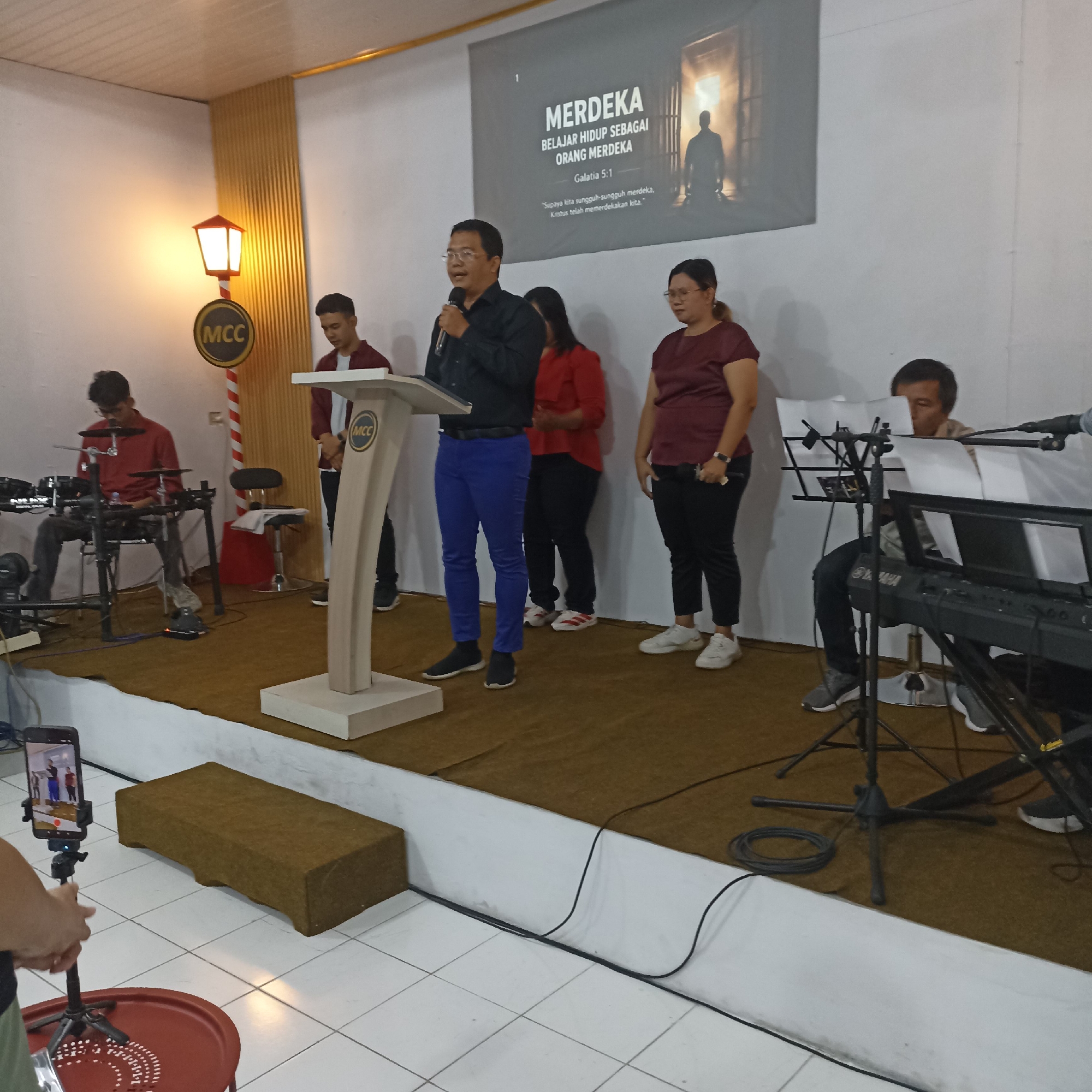 The image shows an indoor gathering, possibly a church service, taking place on a raised platform. A man in a black shirt and blue pants is speaking into a microphone at a lectern with an "MCC" logo. Behind him, several other people are standing, including a woman in a red shirt and another in a maroon shirt. To the left, a young man in a red shirt is playing electronic drums. To the right, a man is seated at a keyboard. A banner with text in Indonesian that reads "MERDEKA BELAJAR HIDUP SEBAGAI ORANG MERDEKA" and a Bible verse is displayed on the wall behind the speakers. The platform is covered with a brown carpet. In the foreground, a smartphone on a tripod is recording the event. The overall setting appears to be a community or religious center. The presence of the "MCC" logo and the general atmosphere suggest it could be a gathering at a church or similar organization. Without specific visual cues or external information linking this scene to Magelang, Indonesia, it is not possible to confirm the location.