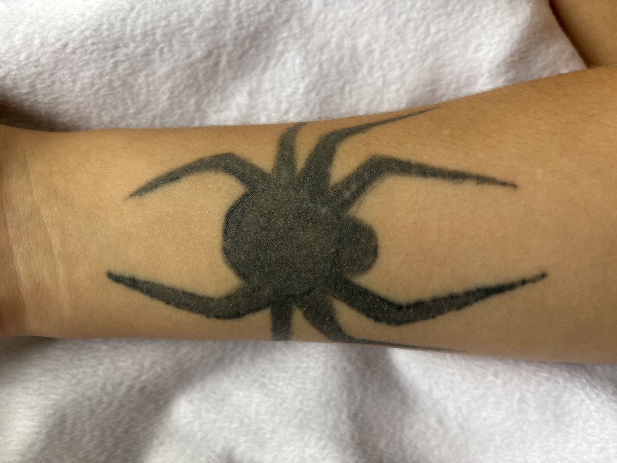 The image displays a black tattoo of a stylized spider on a person's forearm. The spider has a circular body with four legs extending outwards on each side, forming a symmetrical design. The tattoo is situated on skin that appears to be light-toned, with some fine hairs visible. The background of the image is a textured white surface, possibly a towel or blanket. The lighting suggests an indoor setting, with no strong shadows indicating a specific time of day or weather conditions. No text is visible in the image. The overall presentation is a close-up shot focusing solely on the tattoo and the arm it is on.