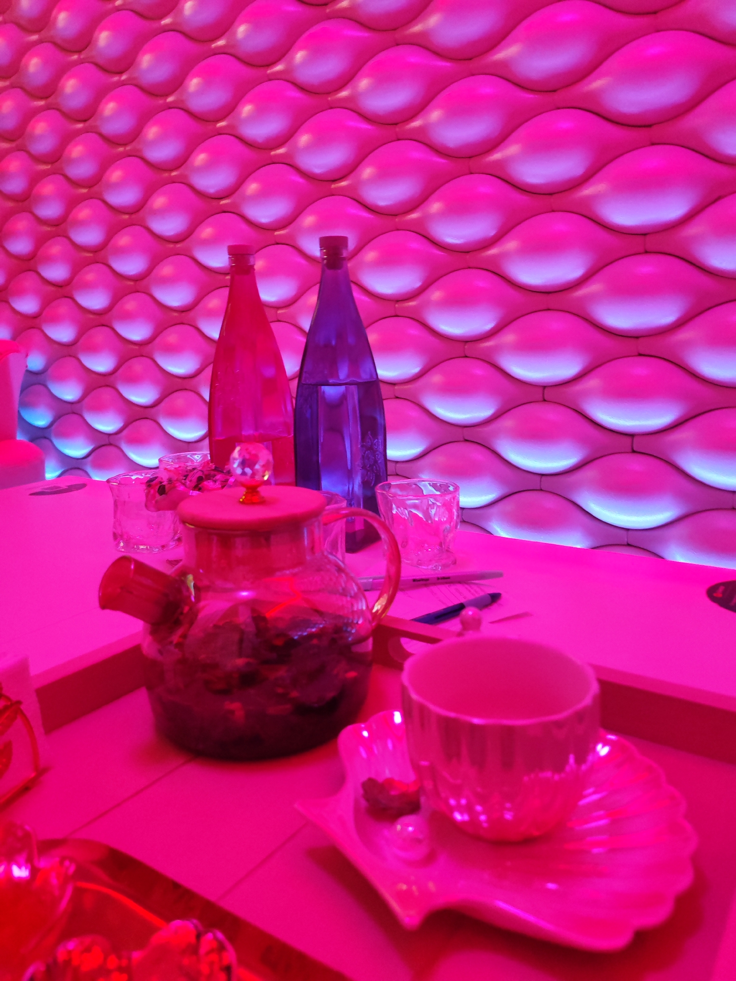 The image captures an indoor setting illuminated predominantly by pink light. The background features a wall with a repetitive, wave-like textured pattern, with embedded lighting creating blue highlights along the lower edges of the waves. In the foreground, a table displays a glass teapot filled with a dark substance, possibly tea, along with two water bottles, one appearing red and the other blue in the pink light. There are also two glasses and a cup and saucer set on the table. Pens and a notebook suggest the table is being used for writing. The overall impression is that of a stylized, possibly luxurious, environment. The location in Izhevsk, Russia is not evident from the image.