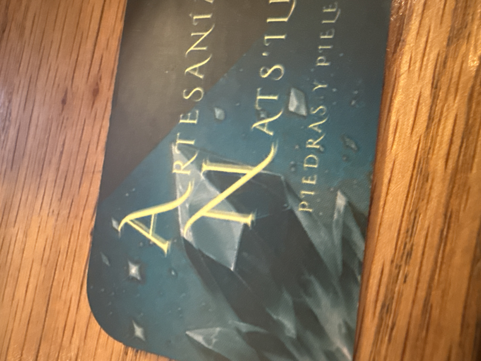 The image shows a business card for "Artesania Xats'il," subtitled "Piedras y Piele." It is photographed on a wood surface. There is no other information.
