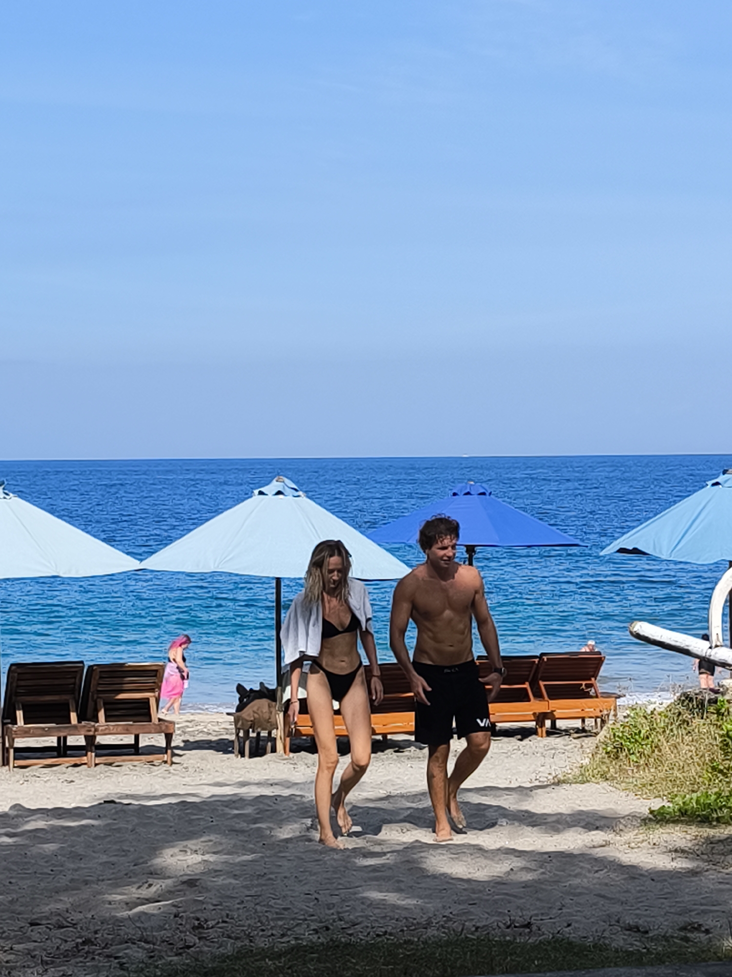 Two individuals, a man and a woman, walk across a sandy beach with the ocean and a clear blue sky in the background. The woman, wearing a black bikini and a white towel draped over her shoulders, walks to the left. The man, shirtless and wearing black swim trunks, walks to the right. In the background, beach chairs and light blue umbrellas are visible, suggesting a resort or public beach area. The ocean is calm and a vibrant blue. The overall scene depicts a serene beach environment. The location is identified as Karangasem, Indonesia.