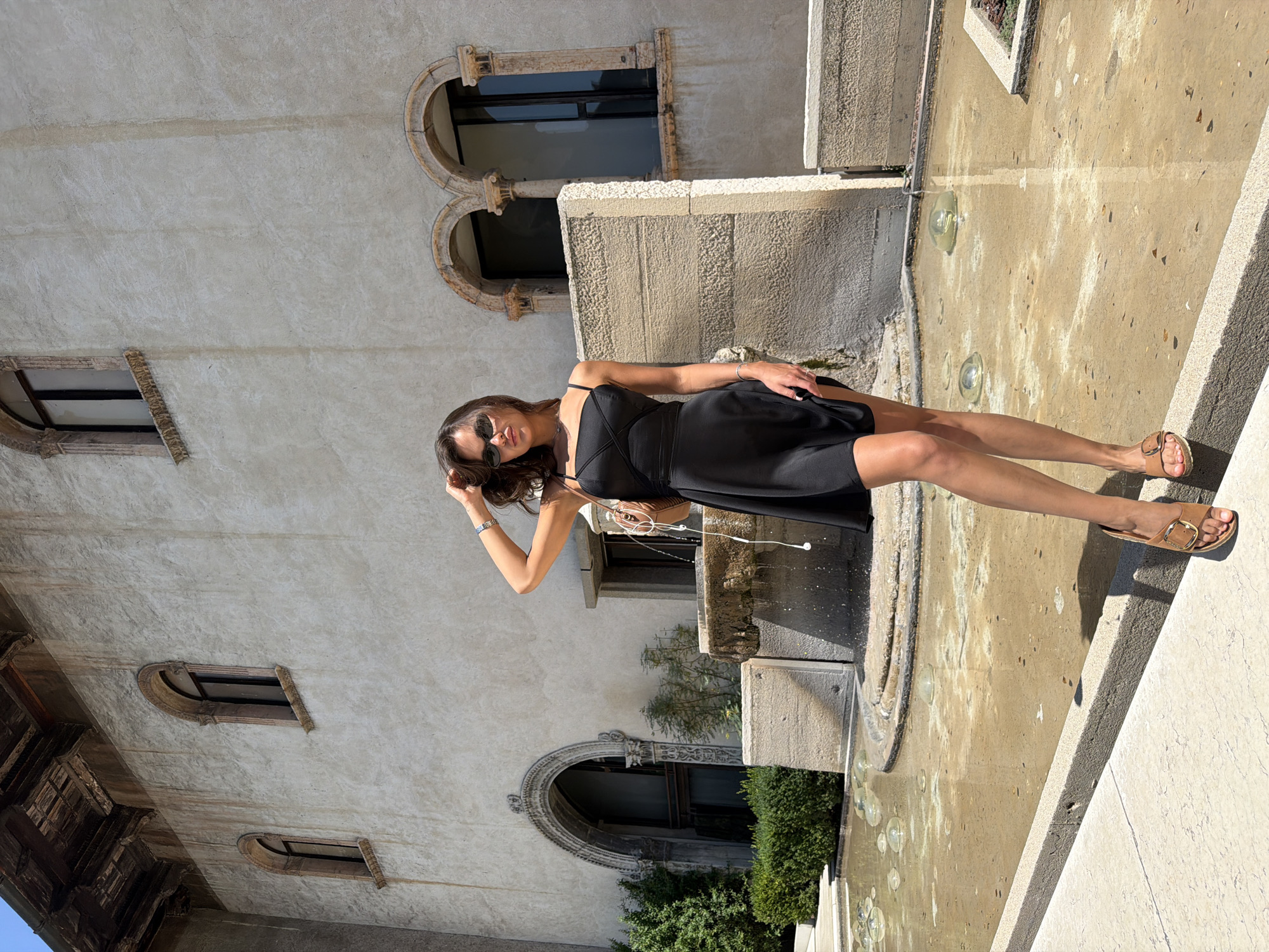 A young woman with dark hair stands in front of a large, aged building with arched windows. She wears a black slip dress, sunglasses, and brown sandals. Her right hand is raised, touching her hair, and she is holding a tan crossbody bag. In the foreground, a shallow fountain with water is visible. The architectural style of the building and the presence of a fountain suggest the location could be in Verona, Italy, a city known for its historical architecture and water features.