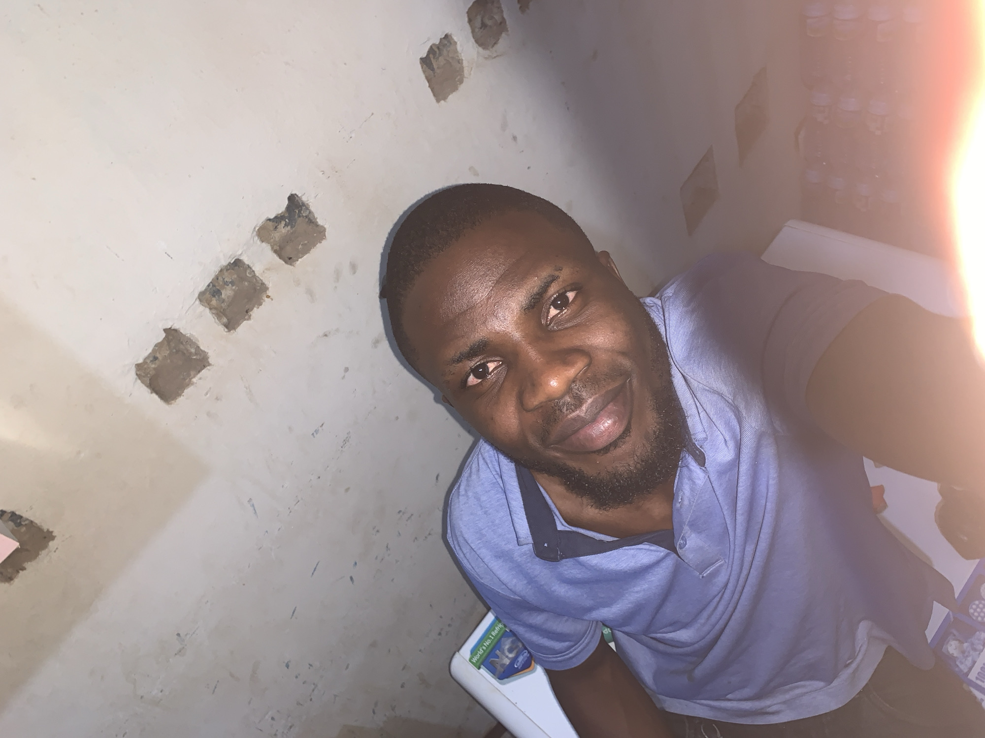 A dimly lit, indoor selfie taken from a high angle features a man with dark skin, a close-cropped haircut, and a neatly trimmed beard and mustache. He is wearing a light blue collared shirt with dark blue trim. The man is smiling slightly, looking directly into the camera.
Behind him is a wall that appears to be unfinished, with several square-shaped holes or indentations revealing the underlayment or bare structure. To the right, slightly behind the man, is a stack of water bottles.
The photograph seems to be taken in a corner of a room, as two walls are visible. A portion of a white object, possibly a piece of furniture or appliance, is visible beneath the man in the frame.
Given the location in the city of New Stadium, Nigeria, no further details can be confidently inferred.