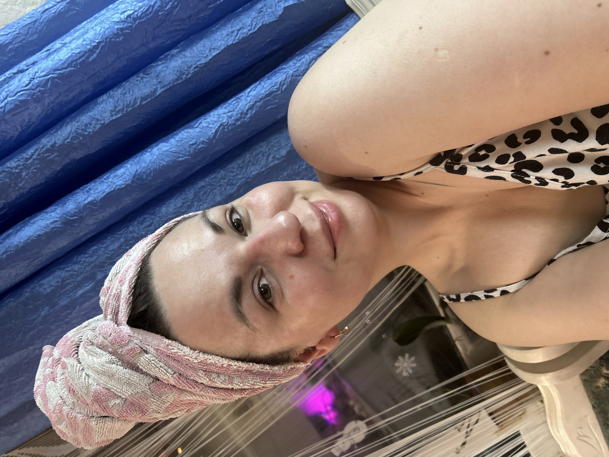 This image depicts a female subject indoors, identified as being in the city of Фоки, Russia. The subject, appearing to be an adult female with fair skin and dark hair, is positioned in a close-up, head-and-shoulders shot, looking directly at the camera with a faint smile. Her hair is wrapped in a pink and white patterned towel, twisted on top of her head. She is wearing a top with a black and white leopard print pattern. A small, golden-colored earring is visible on her left ear. The background on the left features a window partially covered by translucent white vertical blinds or curtains, with a snowflake decal visible on the glass and a blurry purple light source beyond. The right side of the background is occupied by a dark blue, textured curtain hanging in vertical folds.