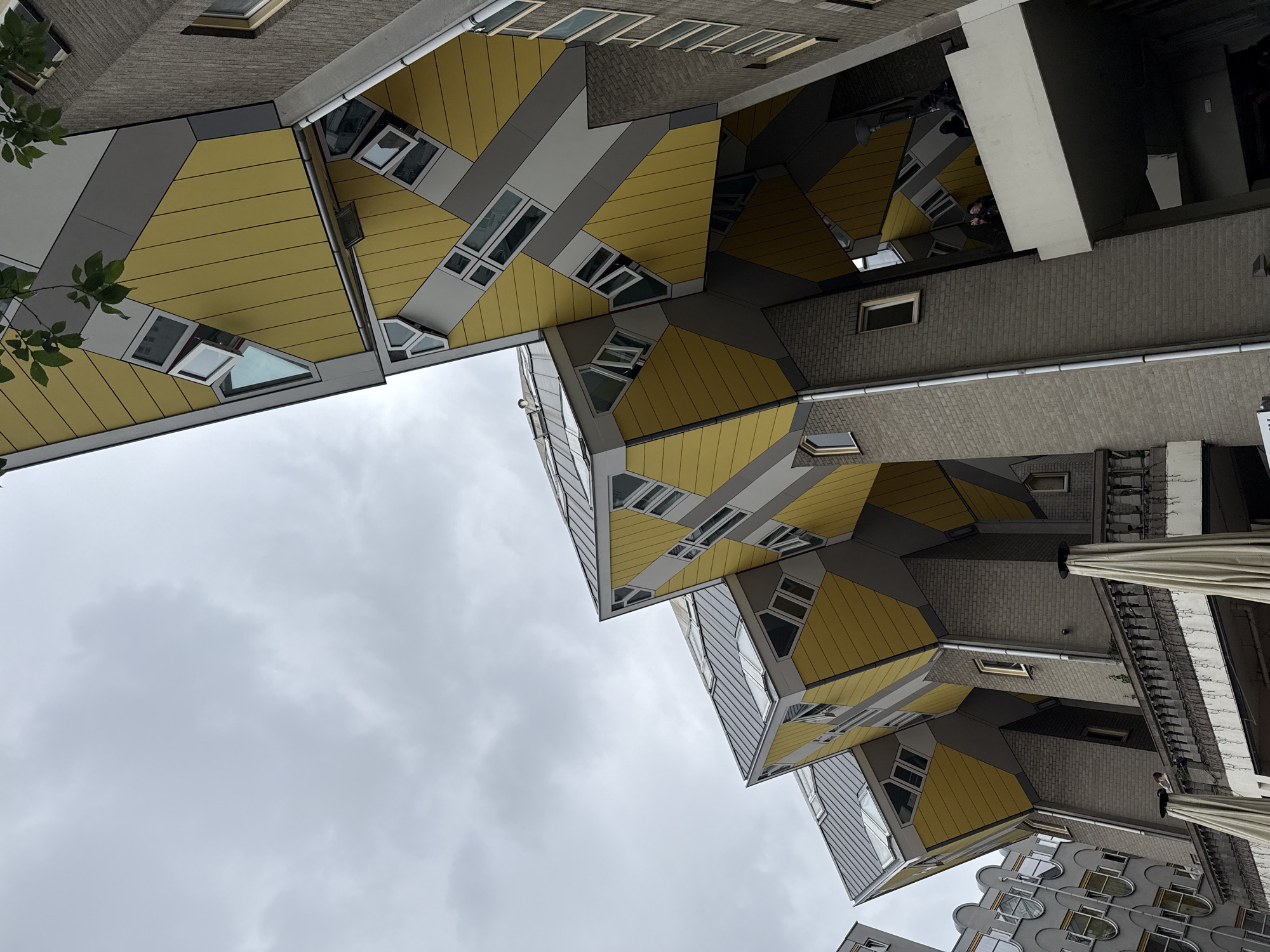 This is a low-angle photograph of the Cube Houses (Kubuswoningen) in Rotterdam, Netherlands, under a cloudy sky. The iconic Dutch architecture features brightly colored yellow, gray, and white geometric shapes that are tilted at sharp angles, creating an abstract, almost surreal cityscape. The buildings are constructed with brick and metal cladding. Windows are visible on many of the tilted surfaces, some open, others closed. In the foreground, a balcony with a railing is partially visible, with two large white umbrellas set up. On the balcony level, two individuals are standing near the railing, appearing to interact with each other and looking towards the camera. The overall impression is one of modern, unconventional design against a somber sky.