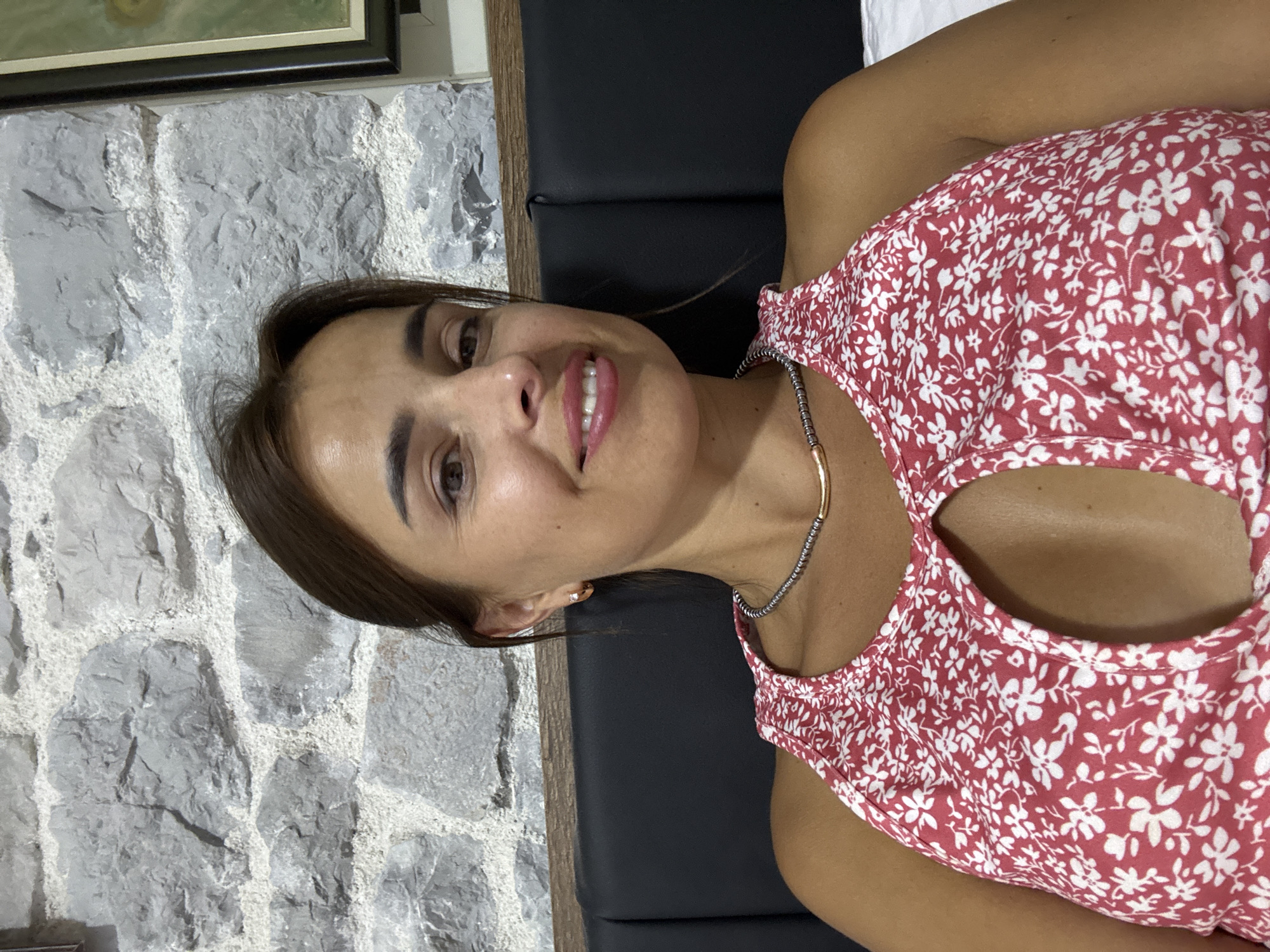 A woman with brown hair and brown eyes smiles at the camera. She is wearing a red and white floral print top with a cutout at the chest and a beaded necklace. She has a small stud earring in her right ear. The background consists of a stone wall and a black headboard.