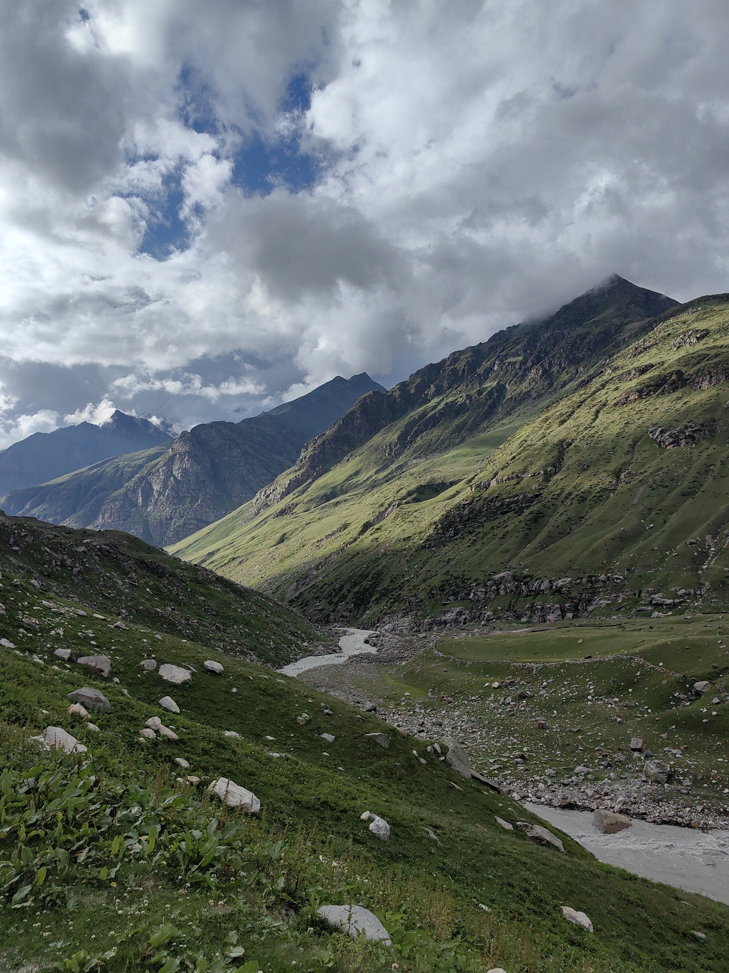 The image depicts a wide, mountainous valley in Burwa, India, under a partly cloudy sky. The valley floor is occupied by a winding, sandy-colored river with some rocks visible in its flow. The slopes of the valley are covered in lush green grass and dotted with scattered rocks and boulders of various sizes. Some areas of the mountainside are illuminated by sunlight, creating a contrast with the shadowed areas. In the distance, successive mountain ranges recede into the background, with some peaks obscured by clouds. The overall scene is a natural landscape characterized by dramatic geological formations and verdant vegetation.