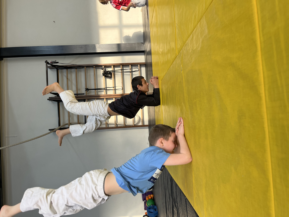 An indoor sports facility in Krasnoyarsk, Russia, features bright yellow interlocking foam mats covering the floor. Two young males are performing forearm stands, inverted with their heads down and hands clasped on the mat. The boy in the foreground, estimated 6-9 years old, wears a light blue t-shirt and white pants. His legs extend vertically upwards, partially obscured by a thick rope hanging from the ceiling. Behind him and to the right, a second boy, estimated 10-14 years old, wears a dark long-sleeved shirt and white pants. He is also in a forearm stand, with his bare feet and lower legs hooked onto the top bar of a wooden wall bar system (stall bars) installed against a light grey wall. A third, younger child, wearing a red t-shirt with a white design and white pants, is partially visible standing upright in the far right background. The room has high ceilings and is illuminated by natural light entering from a source on the right, casting shadows, and likely overhead artificial lighting. Various colorful pieces of gym equipment are visible on a black mat in the far left background. The exact address within Krasnoyarsk cannot be determined from the image.