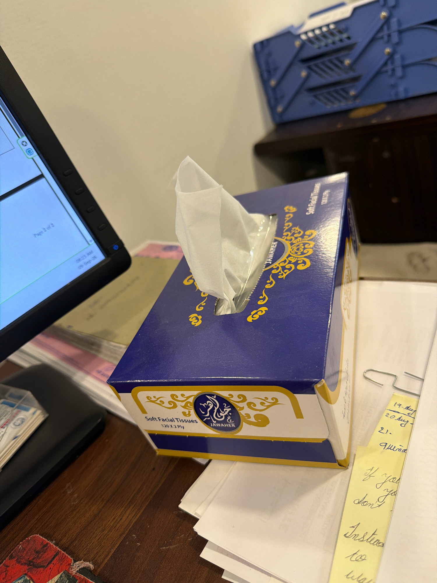 A close-up, high-angle shot depicts a blue and gold box of "JAWAHER Soft Facial Tissues" placed on a wooden desk. The tissue box is partially open, with white tissues protruding from the top. To the left of the tissue box, a portion of a black computer monitor is visible, displaying text that includes "Page 2 of 2". Scattered papers are present on the desk, with a yellow sticky note clipped to one of them. The sticky note contains handwritten notes in blue ink, including dates and what appears to be a message. In the background, a blue plastic organizer with metallic accents rests on a dark surface, possibly a shelf or cabinet. The scene is indoors, likely an office or home workspace, and is located in Riffa, Bahrain.