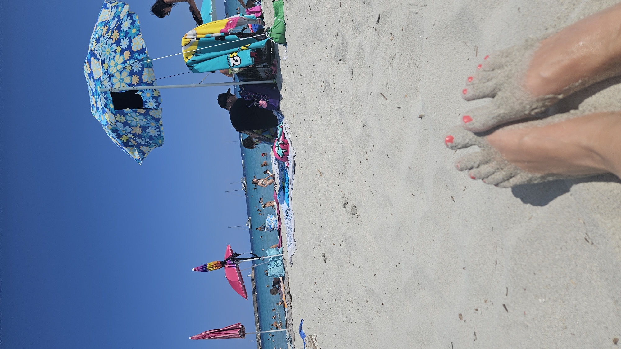 The image depicts a sunny beach scene in L'Île-Rousse, France, focusing on a person's feet partially covered in sand in the foreground. The sandy beach stretches towards the turquoise Mediterranean Sea where numerous people are swimming and relaxing. Several beach umbrellas, one with a blue floral pattern and another pink, are visible, providing shade. Towels and beach bags are scattered on the sand. Boats are anchored in the calm waters. The sky is clear and blue, indicating a warm, pleasant day.