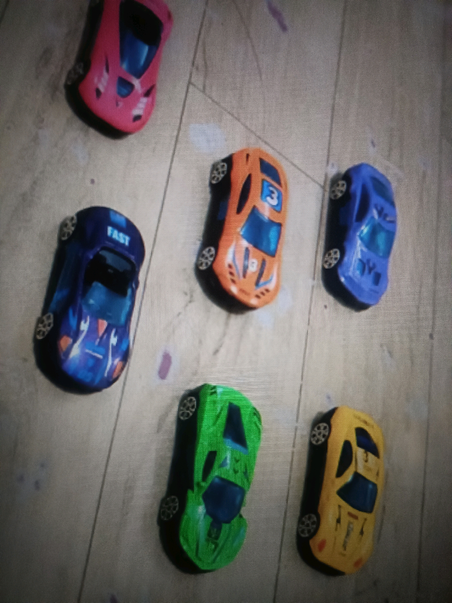 The image shows six toy cars arranged on a light-colored, possibly wooden, floor. The cars are all of a similar size and shape, designed to resemble sports cars. They are positioned in a scattered arrangement, with some overlapping.
Starting from the top left and moving clockwise, the cars are:
1. A pink toy car with a blue windshield and black wheels.
2. An orange toy car with blue accents, a blue windshield, and the number "3" visible on its hood. It has black wheels.
3. A blue toy car with a black windshield and black wheels.
4. A yellow toy car with black accents, a black windshield, and black wheels.
5. A green toy car with blue accents, a blue windshield, and black wheels.
6. A dark blue toy car with "FAST" written on it in white letters. It has blue accents, a black windshield, and black wheels.
The floor has a textured appearance, with visible lines suggesting planks or grout. There are a few faint, light purple smudges on the floor scattered around the cars. The lighting in the image is somewhat dim, creating a slight shadow effect around the objects. The perspective is from directly above, looking down at the cars. There are no people or animals visible in the image.