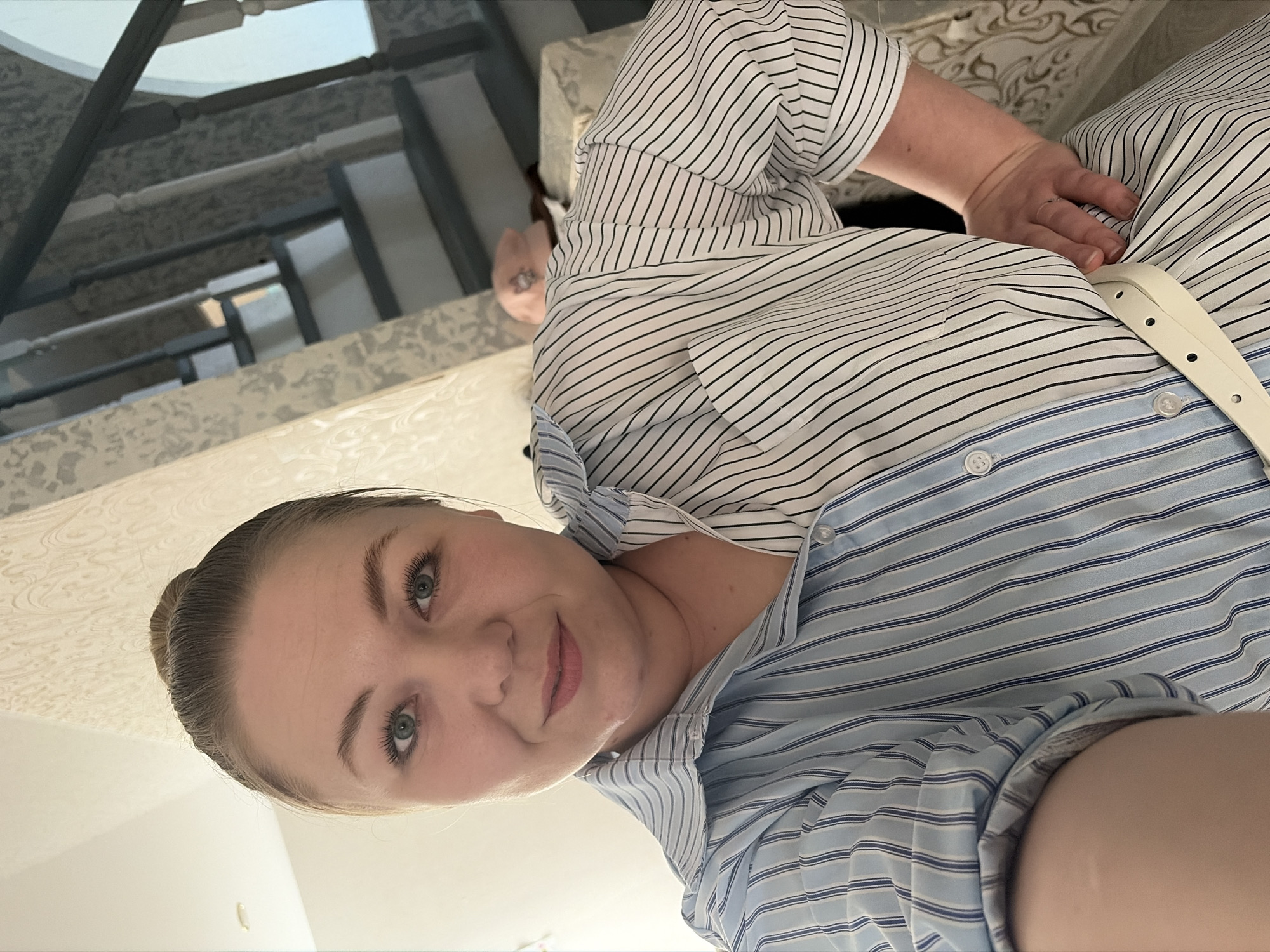 A selfie of a woman with light skin and blonde hair pulled back in a bun, wearing a striped, button-down shirt with a white belt. The environment includes a staircase and furniture, suggesting the interior of a home. There is no indication that the location is Noviy, Russia.