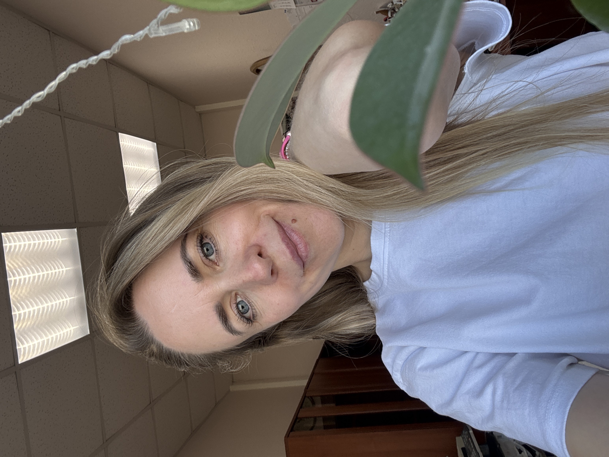 A woman with blonde hair and blue eyes is positioned in the foreground, wearing a white t-shirt. A green plant leaf obscures part of her face. Overhead, there are fluorescent lights in a paneled ceiling. In the background, there is furniture with dark wood. The location is Chaikovsky, Russia.