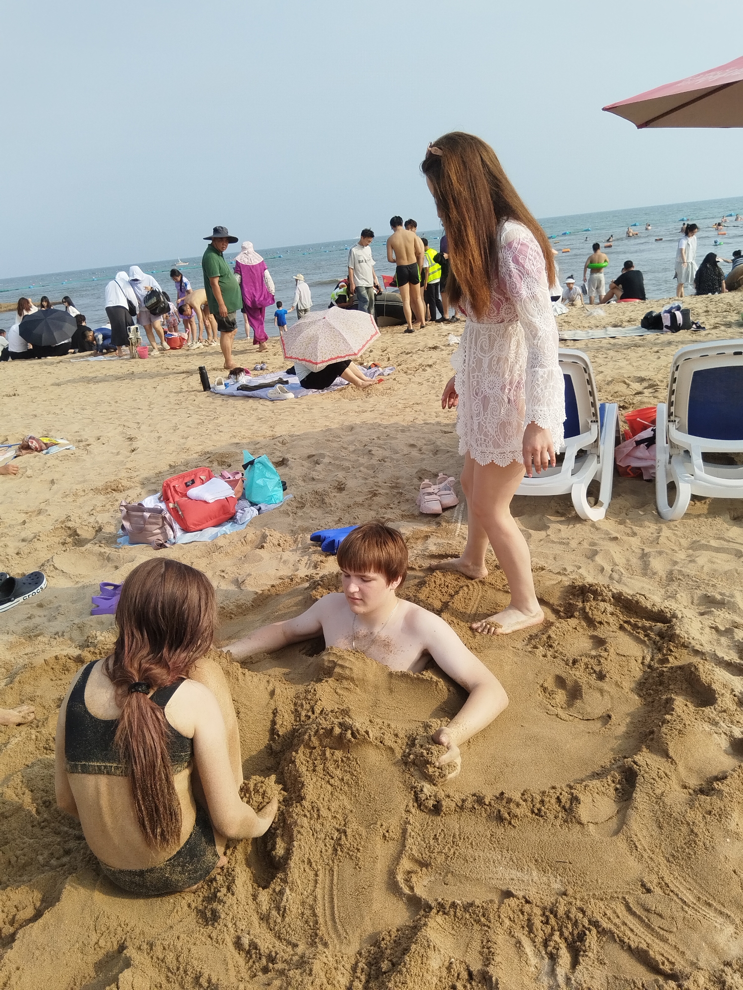 The image depicts a busy beach scene with numerous people enjoying a sunny day at Qinhuangdao, China. The main focus is on a group of individuals engaged in building sand sculptures and playing in the sand. Two young people, a boy and a girl, are partially buried in the sand, appearing to be having fun. The boy has his head and upper body exposed, while the girl's back is visible, with her head turned away from the camera. A woman in a white lace dress stands behind them, observing.
In the background, the beach is populated with many other beachgoers. Some are sitting or lying on the sand, while others are wading in the ocean. There are beach chairs, umbrellas, and various bags and personal belongings scattered on the sand. The sea is calm with a few people in the water, some with inflatable rings.
The time of day appears to be daytime, likely mid-morning to late afternoon, given the bright sunlight and clear sky. The weather is sunny and warm, typical for a beach day. The overall mood seems cheerful and relaxed, with people enjoying recreational activities.
Visible objects include beach chairs, umbrellas, bags (one red, one blue, and one beige), shoes (a black Crocs shoe is visible in the foreground), and inflatable rings in the water. A pink bow is in the hair of the woman in the white dress.
There is no visible text in the image, other than the brand name "Crocs" on the shoe. The location is identified as Qinhuangdao, China.