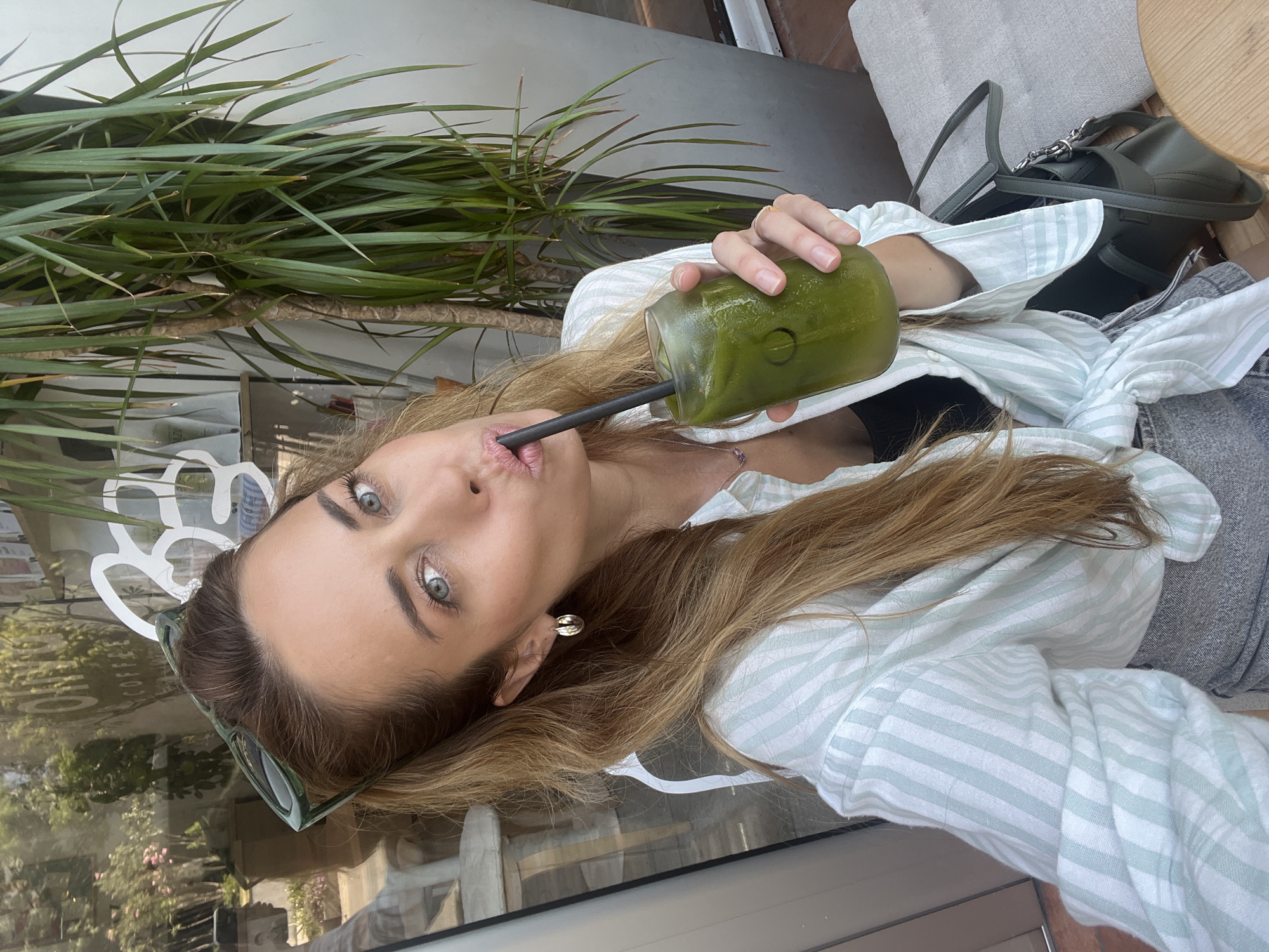 A woman with long, light brown hair is taking a selfie in front of a glass-fronted building. She is wearing a light blue and white striped shirt tied at the waist, a black top underneath, and gray shorts. Her sunglasses are perched on her head. She is holding a glass of green juice with a black straw and is drinking from it. Her left hand has a gold ring on one finger. She is wearing gold hoop earrings. The background shows a reflection of trees and signage that appears to say "COFFEE" and possibly "CO" on a shelf. A large potted plant with long green leaves is to her right. The setting appears to be an outdoor cafe in Bar, Montenegro, given the context provided.
