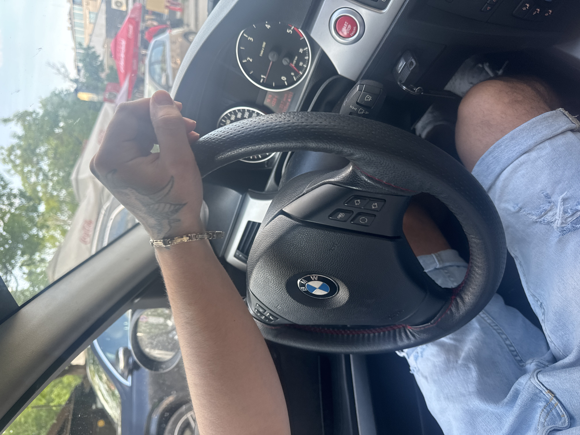 A person is driving a BMW in Sofia, Bulgaria. The driver is wearing light blue ripped jeans and a gold bracelet on their left wrist. The driver's left hand is gripping the steering wheel, and a tattoo is visible on the back of their hand. The car's dashboard and steering wheel are visible. Outside the car window, other vehicles and buildings can be seen. A red Coca-Cola advertisement is visible outside the car window.