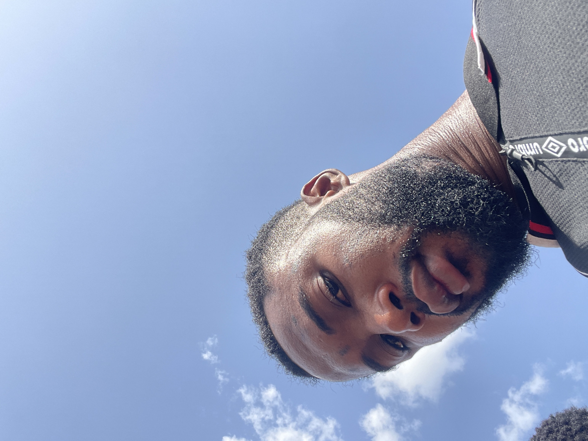 A low-angle shot captures a man with dark skin and a well-groomed black beard against a backdrop of a bright blue sky with scattered white clouds. The man's face is close to the camera, showing sweat on his forehead and around his hairline. He wears a black athletic garment with white and red accents on the upper right. A strap with the Umbro logo is visible on his right shoulder. The specific location in Lokoja, Nigeria, is not discernible from the image.