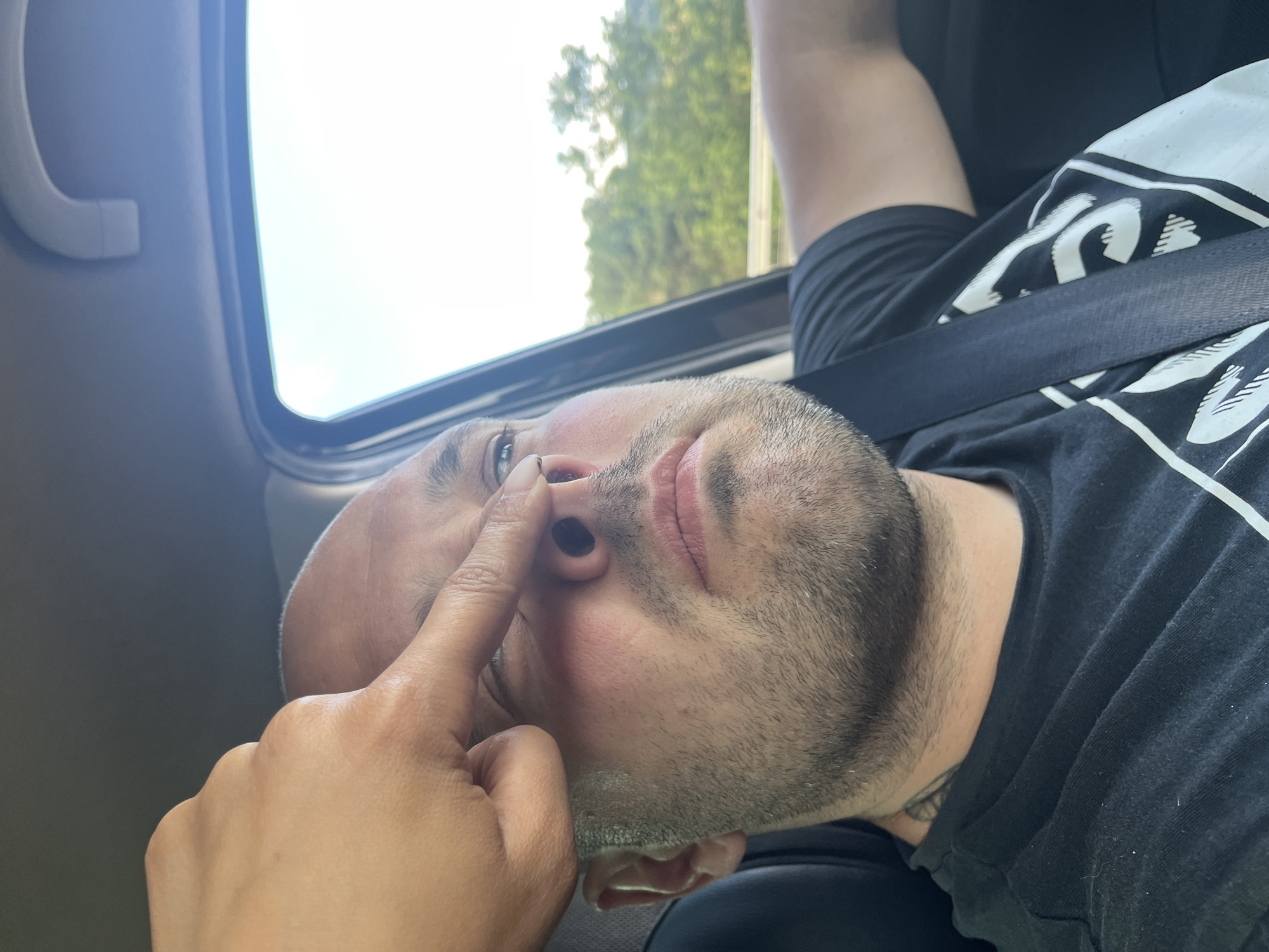 A close-up shot shows a man's face as a finger is pressed against his nose while he sits in the driver's seat of a car. The man is bald, with stubble on his face and a tattoo visible on his neck. He is wearing a black t-shirt with a white graphic design and a seatbelt. The background is blurred, showing trees and a bright sky, suggesting the car is in motion on a road, potentially near Rajana, Bulgaria. The lighting indicates it is daytime.