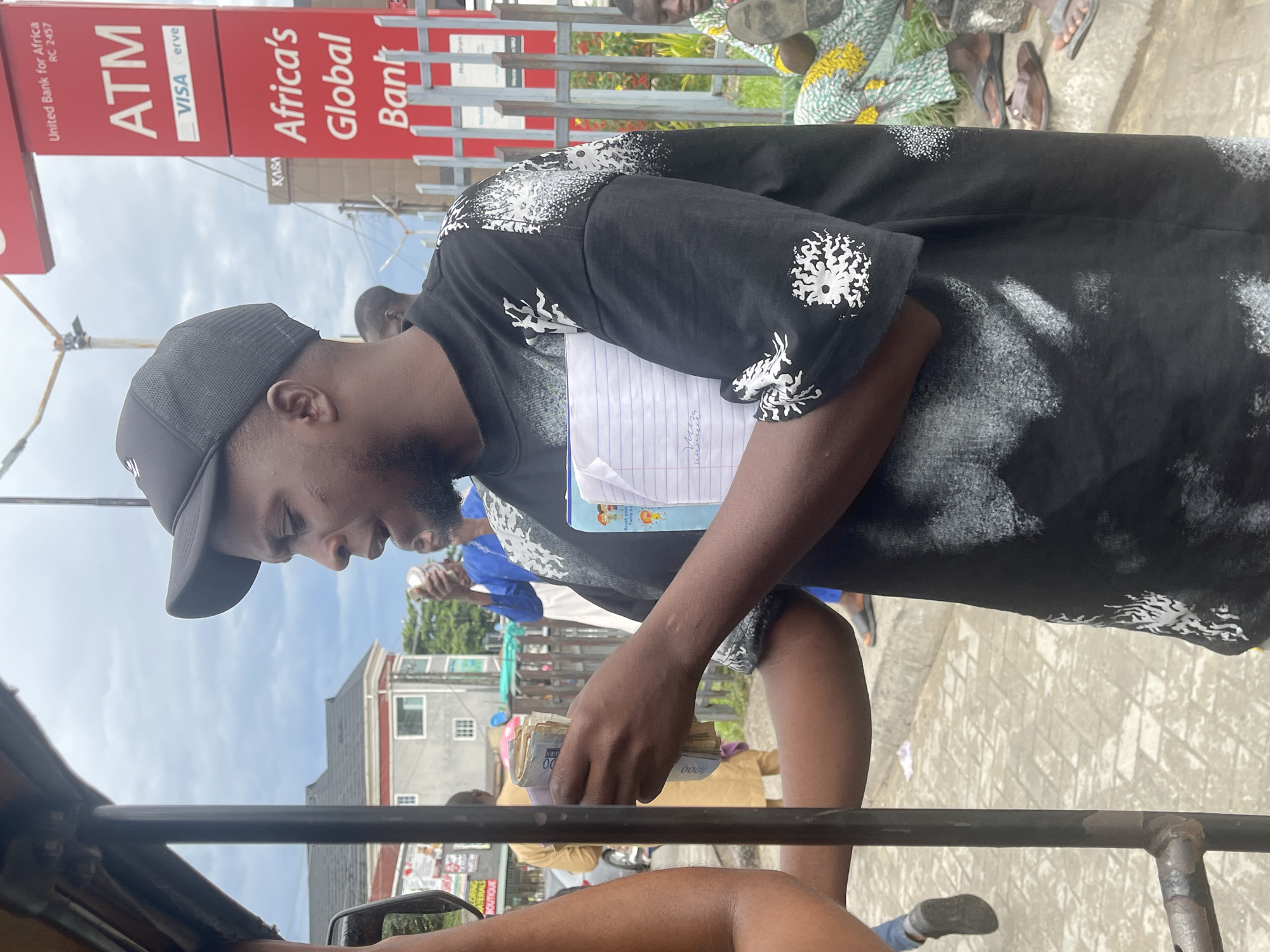 The photo depicts a man counting a stack of Nigerian Naira currency. He is wearing a black t-shirt with white designs, a black baseball cap, and appears to be sitting in some sort of vehicle, possibly an auto-rickshaw or tricycle, based on the visible metal frame. He is holding a notebook with a blue cover on his arm. In the background is a street scene including a red and white ATM sign for United Bank for Africa (UBA) and Africa's Global Bank. Other people are in the background. Given the UBA bank presence and general layout, the location is Ado, Nigeria.