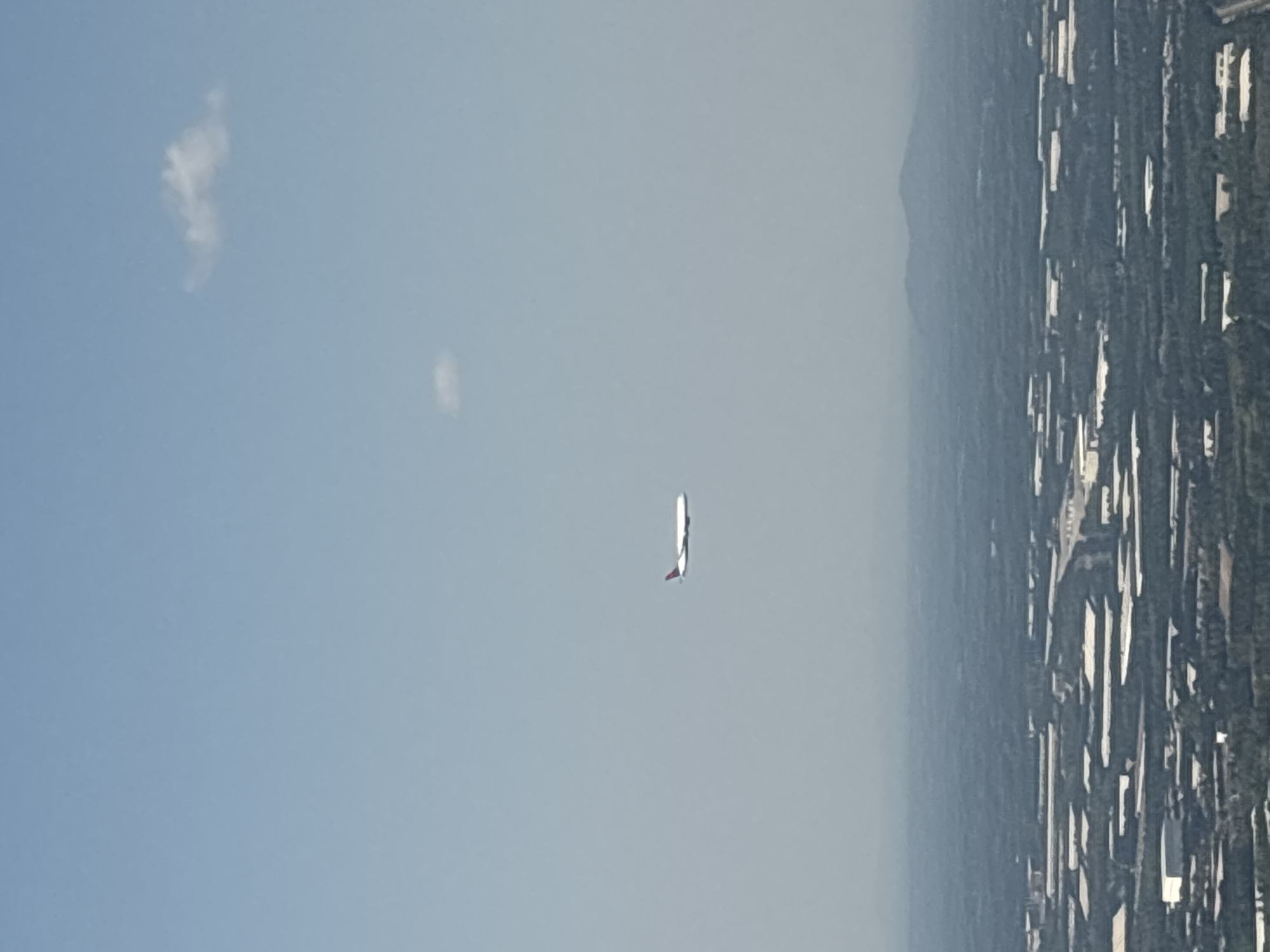 The image shows an airplane flying in a clear sky. In the background is a dense urban area with tall buildings and a mountain range in the distance. There are also a few clouds in the sky.