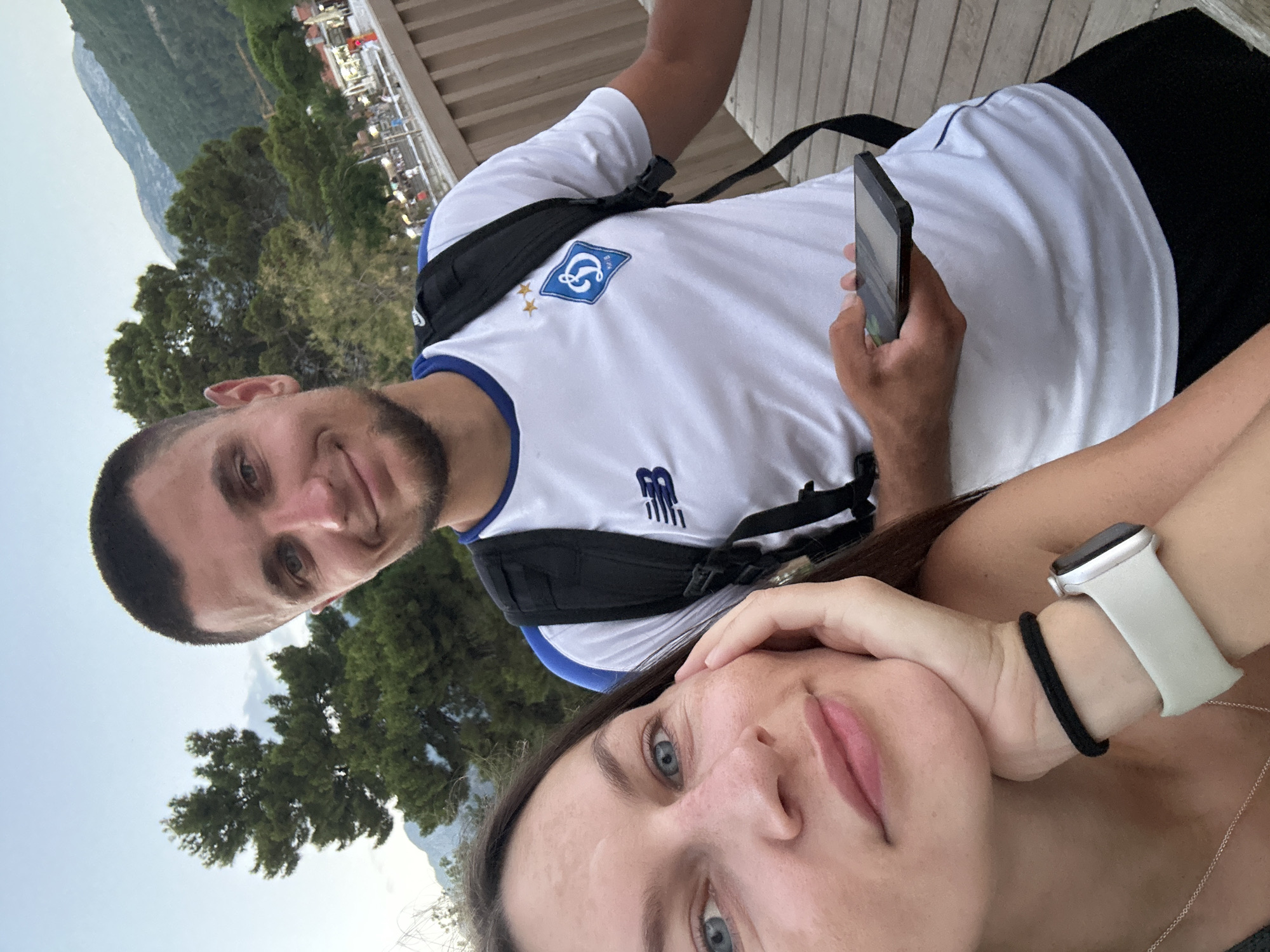 A young couple is taking a selfie outdoors. The woman is in the foreground, with her left hand resting on her cheek and her right hand visible with a smartwatch and a black hair tie on her wrist. She is looking at the camera. Behind her, the man is smiling and looking towards the camera as well. He is wearing a white t-shirt with a blue logo and two yellow stars on the chest, a black backpack, and is holding a smartphone in his right hand. They are standing on a wooden deck. In the background, there are green trees, mountains, and buildings that appear to be part of the city of Budva, Montenegro. The sky is a pale blue.