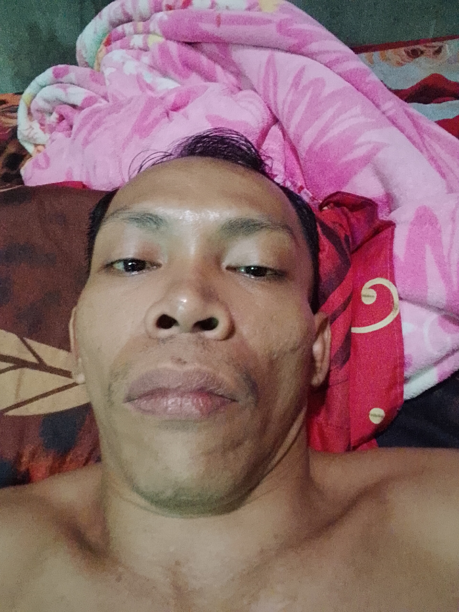 A close-up, selfie-style photograph shows a person lying down with their head close to the camera. The person's face is the primary focus, with their eyes open and looking directly at the lens. Their skin appears somewhat oily, and there are some minor blemishes visible. Their hair is dark and styled upward. They have a bare chest visible below their face. The background consists of a patterned blanket, primarily in shades of pink and red, with some swirls and floral designs. The lower portion of the image shows more of the blanket and some of the person's shoulders and upper chest. The lighting is direct, creating some highlights on the person's forehead and nose. The overall impression is that of a casual, personal photograph taken indoors. No specific landmarks or elements indicating the city of Madukoro, Indonesia are present in the image.
