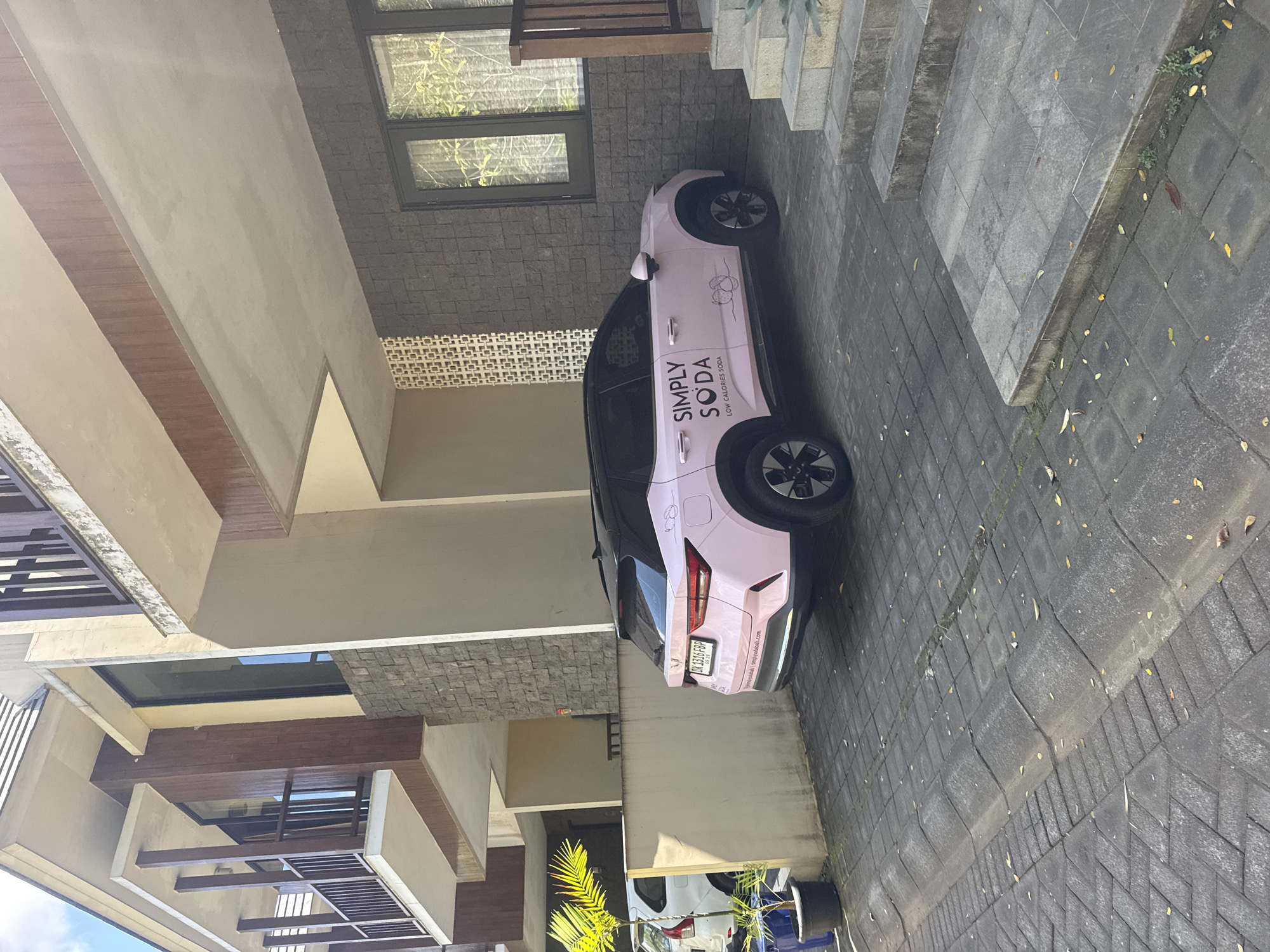 In Kuta Selatan, Indonesia, a light pink electric vehicle, branded with "SIMPLY SODA LOW CALORIES SODA" and a logo of two peas, is parked on a paved driveway in front of a modern multi-story building. The building features a combination of beige stucco, dark stone cladding, and dark wood accents, with balconies on the upper floors. To the left of the vehicle, a white SUV is partially visible, parked next to a potted palm tree. The driveway is made of dark interlocking pavers, with fallen leaves scattered across the surface. To the right of the vehicle, concrete stairs lead up to the building entrance. The scene is lit by natural sunlight, casting shadows on the ground.