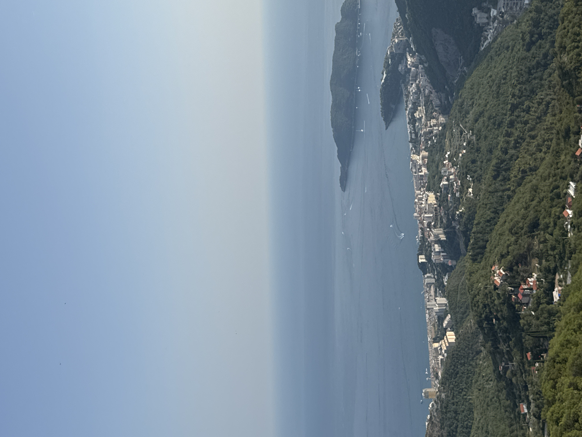 This media file depicts an aerial view of a coastal town nestled on a steep, forested mountainside overlooking a vast expanse of deep blue water. The scene is dominated by the juxtaposition of the lush green of the vegetation and the built-up structures of the town clinging to the slopes. The town itself appears to be a densely packed collection of buildings, ranging from multi-story white structures to smaller residential houses with red-tiled roofs, many of which are partially obscured by the dense foliage.
The water stretches out towards the horizon, where it meets a hazy, pale blue sky, suggesting a distant or slightly overcast day. The overall atmosphere is serene and captures a sense of picturesque tranquility. Several small boats are visible on the water, creating subtle trails and adding a touch of movement to the otherwise still scene. The mountainous terrain is heavily wooded, with the trees densely covering the slopes, creating a textured green carpet that contrasts with the architectural elements of the town.
The perspective suggests the image was taken from a high vantage point, possibly from a drone or a very high altitude. The lighting indicates it is daytime, with the sun casting a soft illumination that doesn't create harsh shadows, likely mid-morning or late afternoon. The location cues, such as the dramatic mountainous coastline and the style of the architecture, are consistent with the context of Brajici, Montenegro, a region known for its stunning Adriatic coastal scenery and steep, verdant hills. There is no visible text in the image.