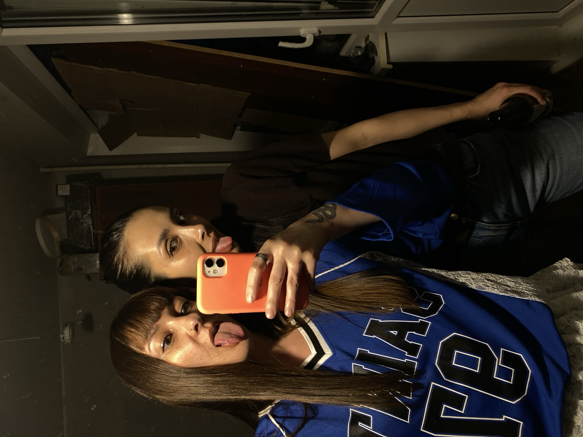A playful mirror selfie captures two young women indoors in Votkinsk, Russia, under dim lighting conditions suggesting evening or low ambient light. In the foreground, a woman with long dark brown hair and bangs is holding an orange smartphone, presumably to take the photo. She is wearing a blue sports jersey with white and black lettering that reads "LA" and "FE 78", and is sticking out her tongue in a humorous gesture. Behind her, another woman with dark hair pulled back is visible, wearing a dark brown t-shirt and blue jeans. She has a snake tattoo on her left forearm and is holding a dark bottle, possibly a drink. She is also sticking out her tongue, mirroring the playful expression of the other woman. The background features indistinct elements such as a white window frame and dark wooden or cardboard panels, indicating a casual indoor setting. The overall mood conveyed is relaxed and jovial.