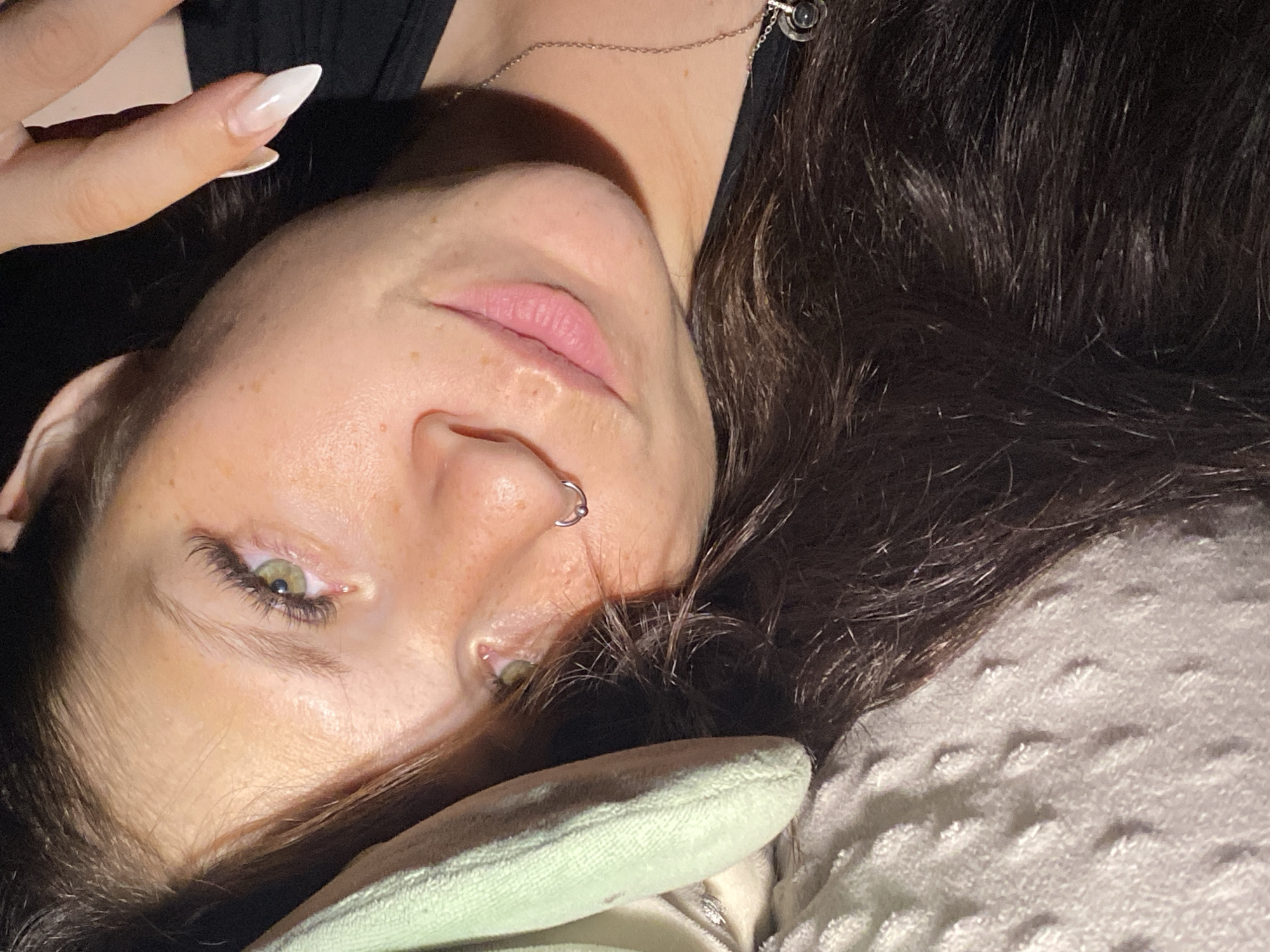 The image is a close-up, downward-angled selfie of a young woman lying on her side, looking up at the camera. She has long, dark brown hair, green eyes, and freckles on her face. She is wearing a nose ring and a necklace. She appears to be indoors, with soft lighting and a textured blanket or pillow beneath her. There is no indication of the city of Chaikovsky, Russia, or any other specific location.