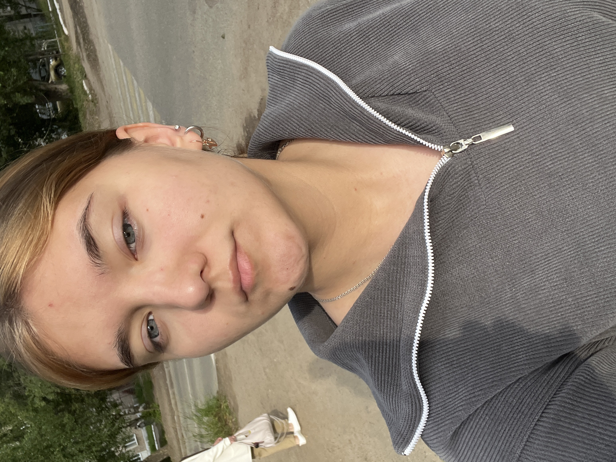A young woman with light brown hair and blue eyes is posing for a selfie outdoors. She is wearing a gray zip-up jacket with a white zipper. In the background, a woman in a cream-colored jacket and tan pants is walking, carrying a light-colored bag. The environment appears to be an urban setting, possibly a street in Chaikovsky, Russia, with a sidewalk and trees visible. The overall lighting suggests it is daytime.