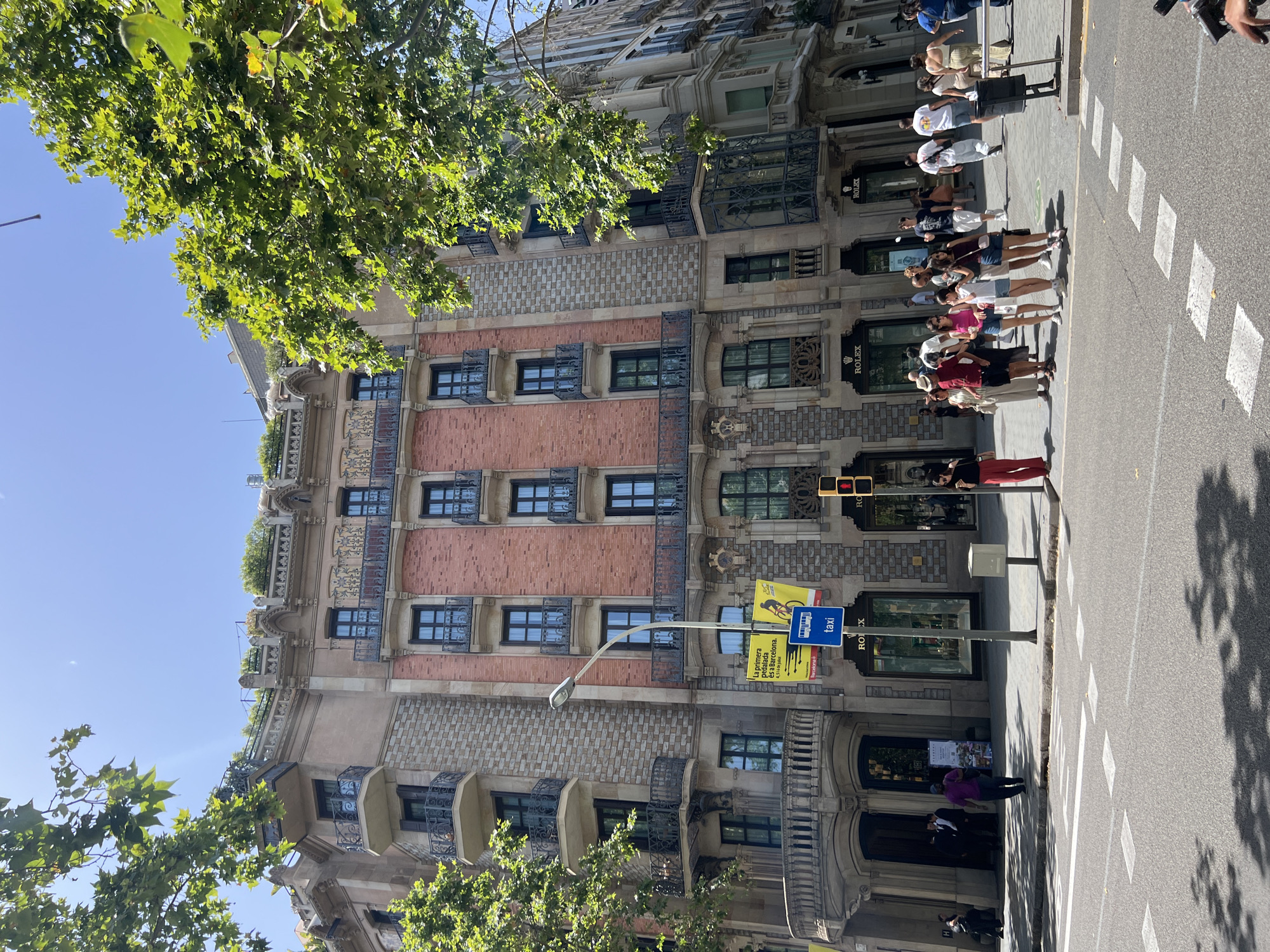 The image shows a street scene in Barcelona, Spain, on a sunny day. A group of people are standing on the sidewalk, some of them are looking at the buildings. The architecture of the building is ornate with brick and stone facades, intricate metalwork balconies, and multiple windows. A large tree with green leaves overhangs the scene from the left.
In the background, people are queuing or walking along the sidewalk. Some of the shops visible have signs for "Rolex." There are also street signs, including a blue sign with "taxi" and yellow signs with graphics and text. One yellow sign reads "La primera... peatonal..." and "...a Barcelona." The street has white pedestrian crossing markings. The time of day appears to be midday, with strong sunlight casting shadows. The overall atmosphere is one of a typical urban environment on a pleasant day.