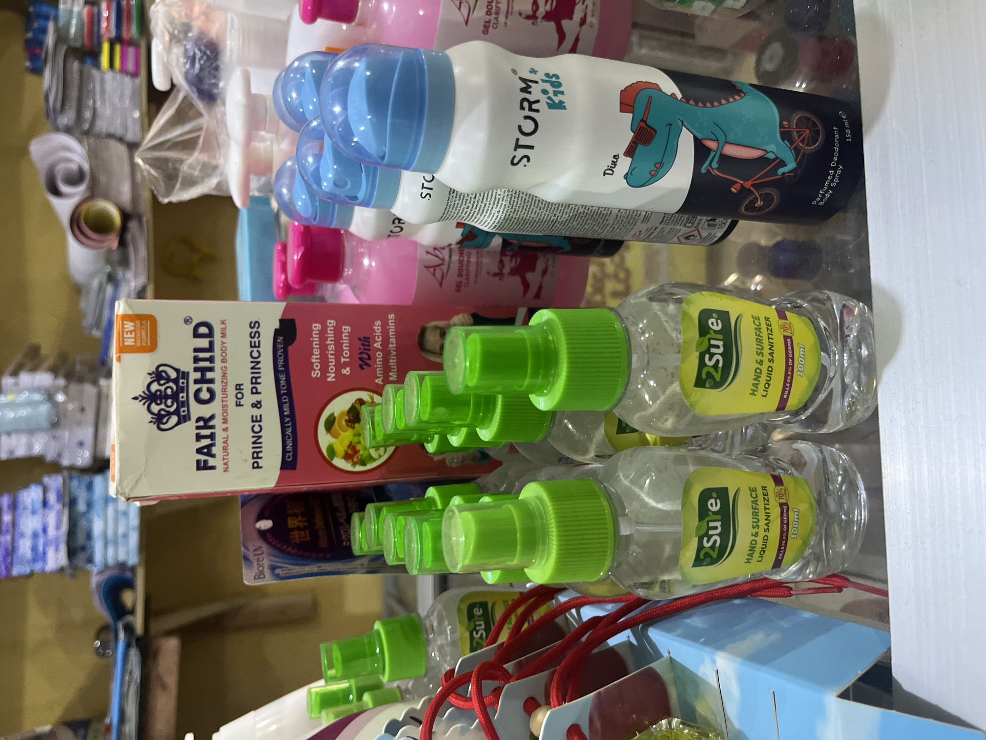 This image displays a retail setting, likely a shop, with various hygiene and personal care products arranged on shelves. The focus is on a collection of hand sanitizers and body milk, with a prominent box of "FAIR CHILD" body milk for "PRINCE & PRINCESS" visible in the foreground. Adjacent to it are several clear plastic bottles of "2Sure" hand and surface liquid sanitizer with green spray nozzles.
Behind these, a row of "STORM * Kids" perfumed deodorant body sprays is stacked. These bottles feature cartoon illustrations of a dinosaur. There are also other indistinct bottles, some appearing to be body wash or similar products, in shades of pink and blue. The overall impression is of a well-stocked display of consumer goods. The presence of items like hand sanitizer suggests a context where hygiene products are readily available, possibly in a local market or convenience store in Bukuru, Nigeria. The lighting appears to be artificial, typical of an indoor retail environment. There are no people visible in the image.