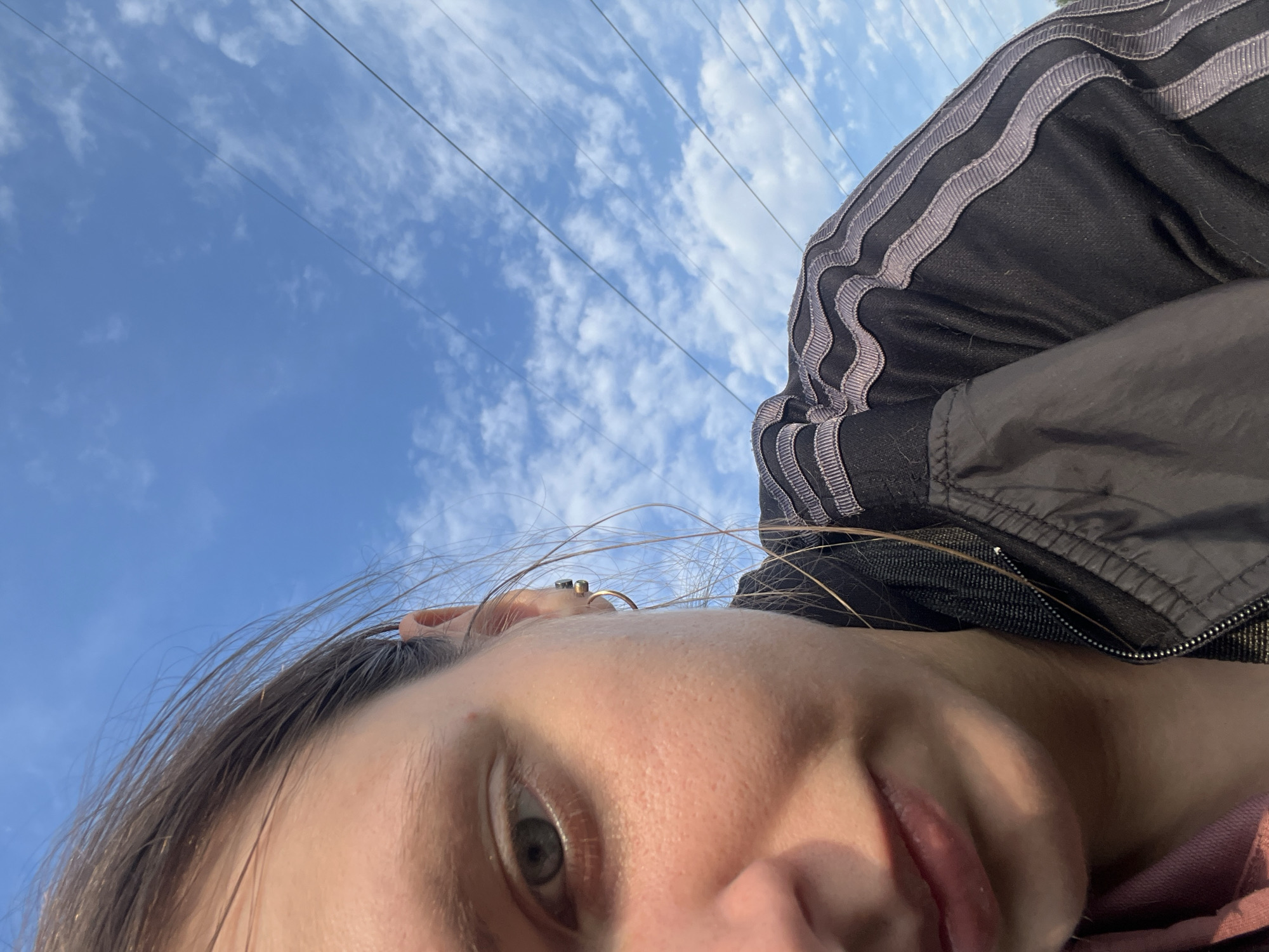 This is a close-up, low-angle selfie of a young woman against a bright blue sky with scattered white clouds. She is wearing a black jacket with distinctive gray Adidas-style stripes on the shoulder and sleeve. The lower part of the frame shows her face, with her eye, nose, and mouth visible. She has brown hair and is wearing earrings, including a gold hoop. Her lips are slightly parted. The image appears to have been taken outdoors on a clear day, likely during daylight hours, given the bright sky and sunlight on her skin. The presence of power lines in the background suggests an outdoor, possibly urban or semi-urban, setting. The overall tone of the image is casual and spontaneous, possibly capturing a moment of self-expression or a lighthearted pose. There is no visible text in the image. The location context is Nizhny Tagil, Russia.