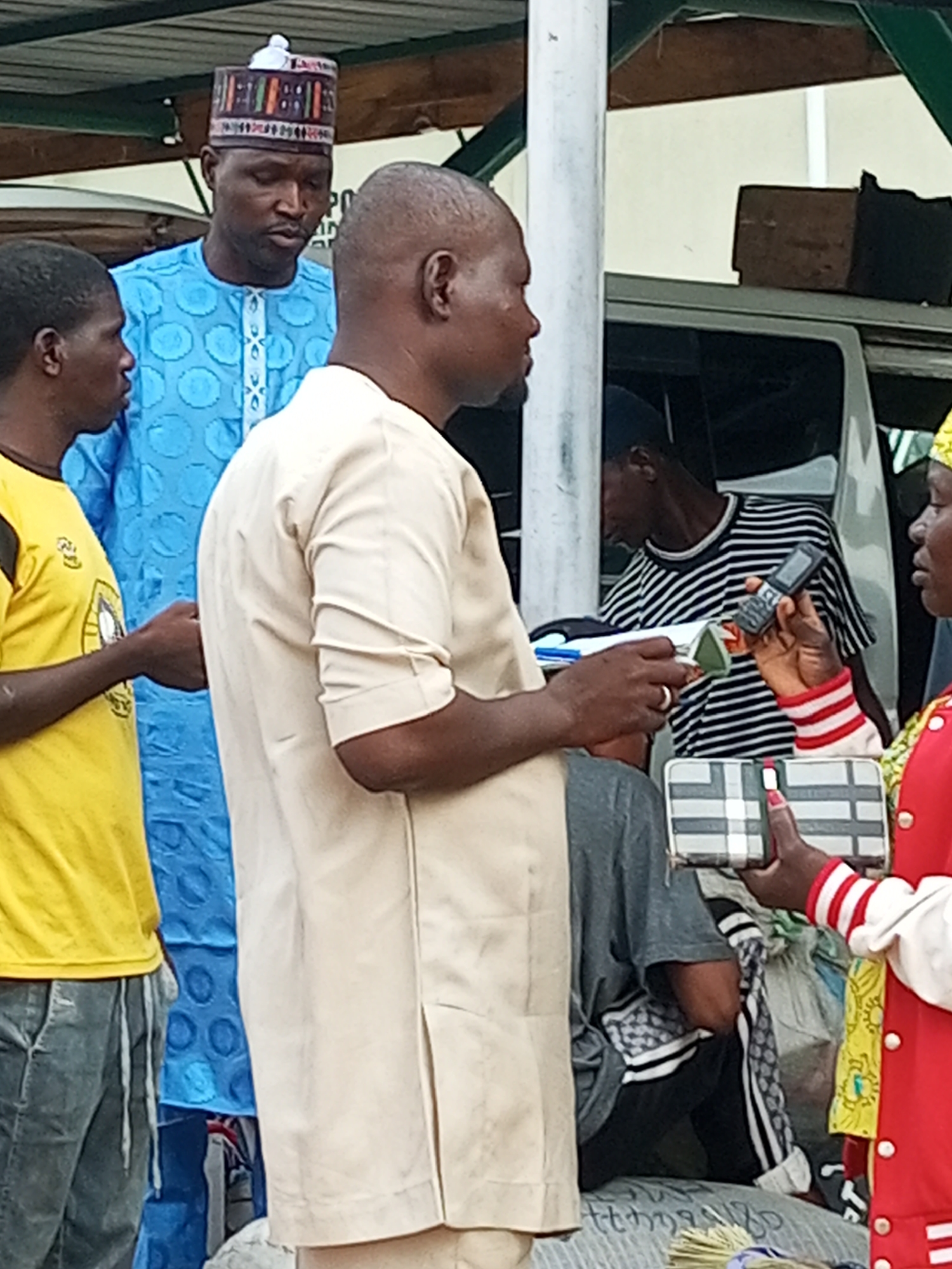 The image depicts a street scene in Gombe, Nigeria, with several individuals present. In the foreground, a man in a light-colored garment is holding a small object, possibly a phone, while interacting with another person whose arm is visible. To the left, a man in a bright yellow t-shirt and grey pants stands facing the center. Behind them, a man wearing a blue traditional outfit and a patterned cap looks down. Further in the background, a person in a striped t-shirt can be seen. The environment suggests an outdoor market or a public gathering area, with a vehicle partially visible and some goods piled up. The lighting indicates it is daytime.