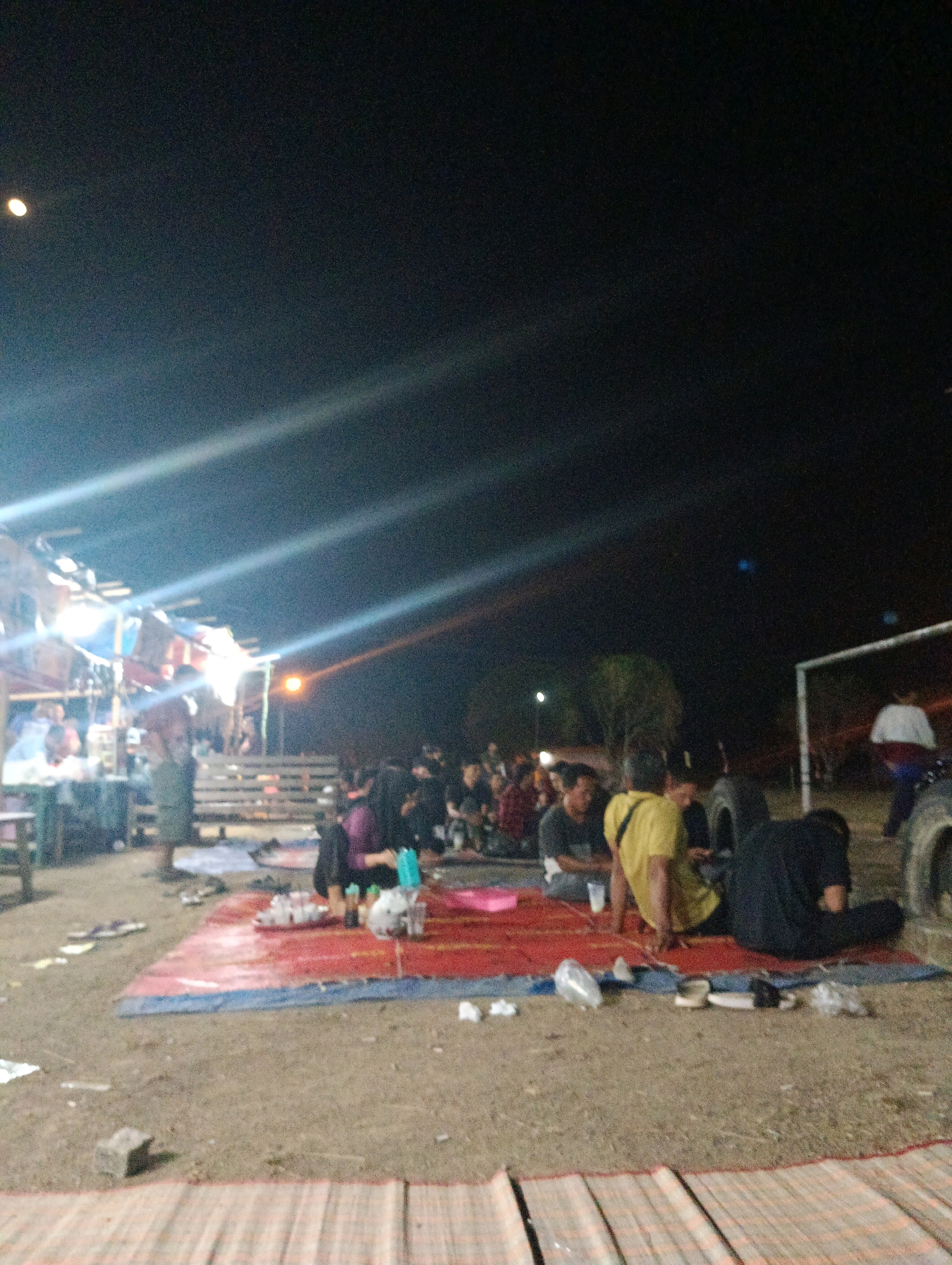 The image shows a group of people gathered outdoors at night, likely in Pematang Jaya, Indonesia. The scene appears to be a casual social gathering, possibly a street food stall or a community event. Several individuals are seated on mats spread on the ground, engaged in conversation or eating. Some are illuminated by bright artificial lights, creating strong glare and lens flares across the dark sky. In the background, there are structures that could be stalls or temporary shelters, with some lighting visible. Further in the distance, a white structure resembling a goalpost suggests an open area or sports field. The overall atmosphere is that of an informal outdoor event taking place after dark.