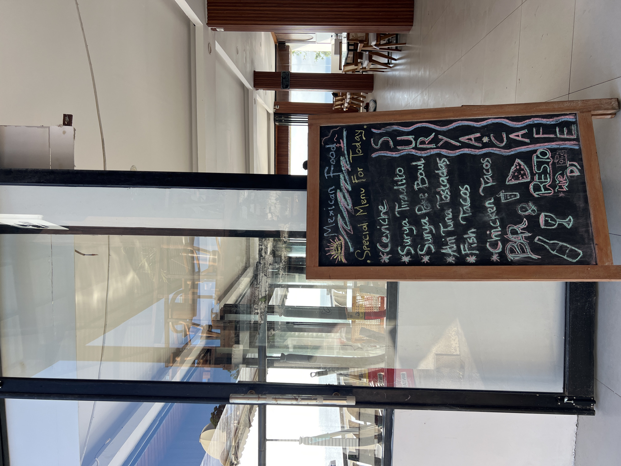 A chalkboard menu stand outside Surya Cafe in Unknown, Unknown lists "Mexican Food" and a "Special Menu For Today" with items like Ceviche, Surya Tiradito, Surya Poke Bowl, Ahi Tuna Tostadas, Fish Tacos, and Chicken Tacos. The sign includes "BAR & RESTO" with sketched images and "Live DJ". The sign is set outside a glass door/window, with glimpses of seating and a possible distant sea view. The location is brightly lit.