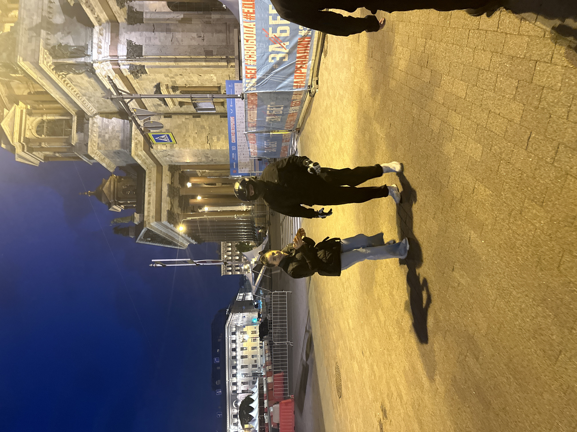 The image shows a street scene in Saint Petersburg, Russia at night. On the left, there is a large building with classical architecture, illuminated by streetlights, and part of Saint Isaac's Cathedral can be seen. The sky is dark blue. On the right, the street is paved with light-colored stones and illuminated by yellowish streetlights. There are two people in the image, one with a motorcycle helmet and black clothing, and the other with a black coat and light-colored pants. A portion of a person's bare foot is visible at the top of the frame. There is a temporary barrier with writing in Russian. There are also multiple buildings visible in the distance.