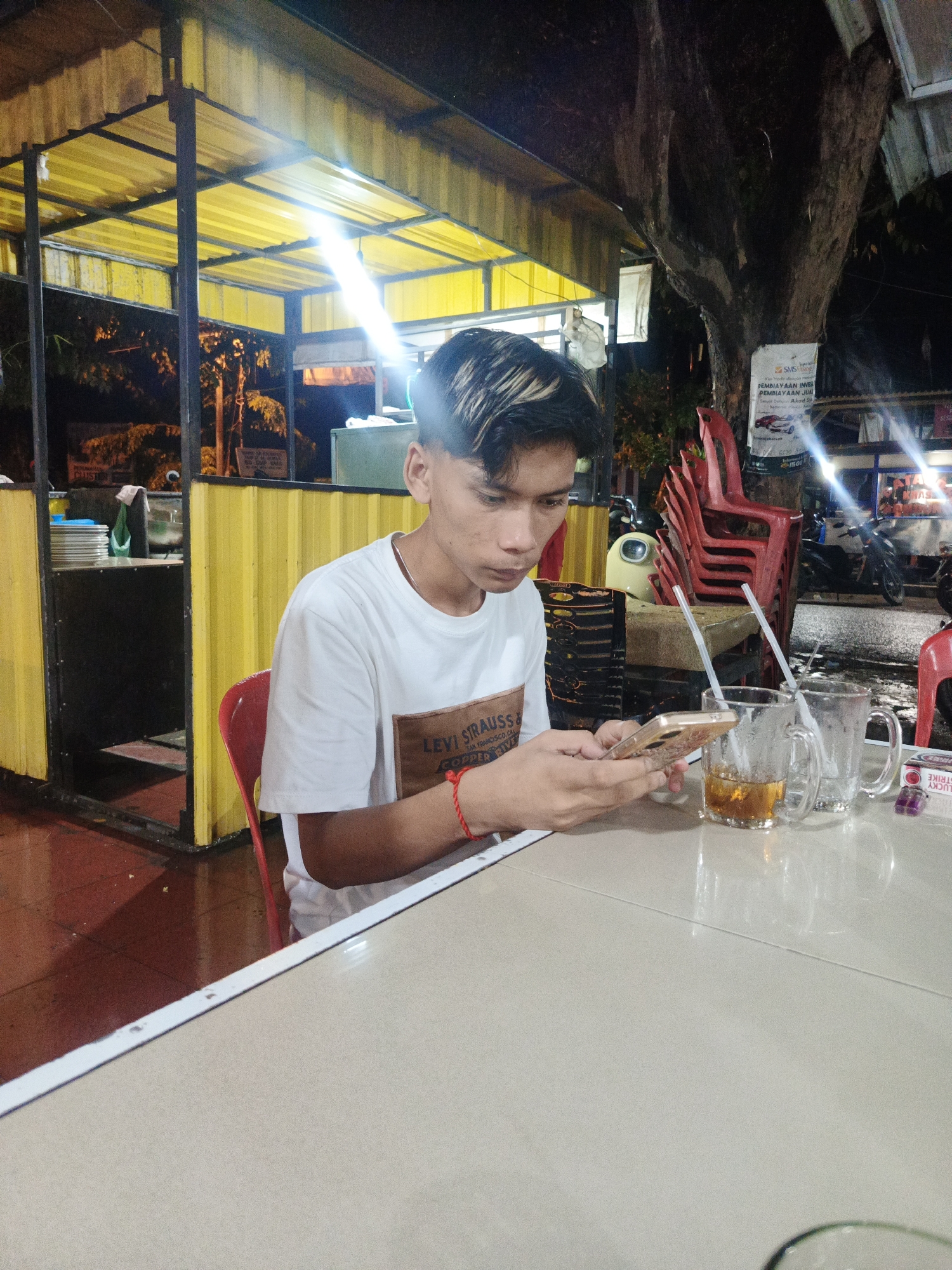A young man is sitting at a table outside, illuminated by overhead lighting, using his smartphone. He is wearing a white t-shirt with a "Levi Strauss" logo, and a red bracelet. There are two glasses on the table, one containing liquid, each with a straw. A pack of "Lucky Strike" cigarettes is also on the table. Red plastic chairs are stacked behind him. In the background, there are motorbikes and buildings, and a sign advertising financial services. Based on the presence of Indonesian text and "Lucky Strike" cigarettes, the location is potentially Lhokseumawe, Indonesia.