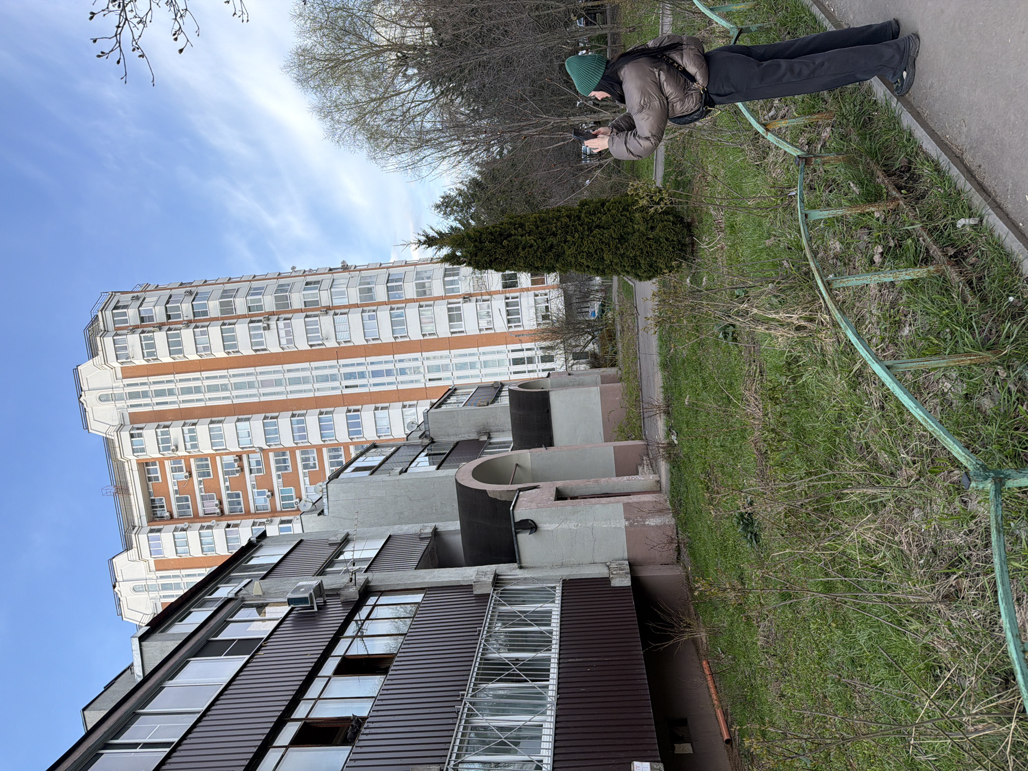 A vertical, eye-level photograph depicts an urban scene, likely in Brekhovo, Russia, showing a building with brown and white facade, a tall apartment complex, a woman using a phone, and surrounding vegetation. The woman is located on a sidewalk adjacent to the building and a green metal fence. The sky is visible in the upper left, showing the blue sky and white clouds. The camera's angle includes a view of a tree, and part of the brown and white apartment building. The other building is taller, and it has an orange and white facade. The picture is taken in the afternoon with the sunlight hitting the buildings and the vegetation.