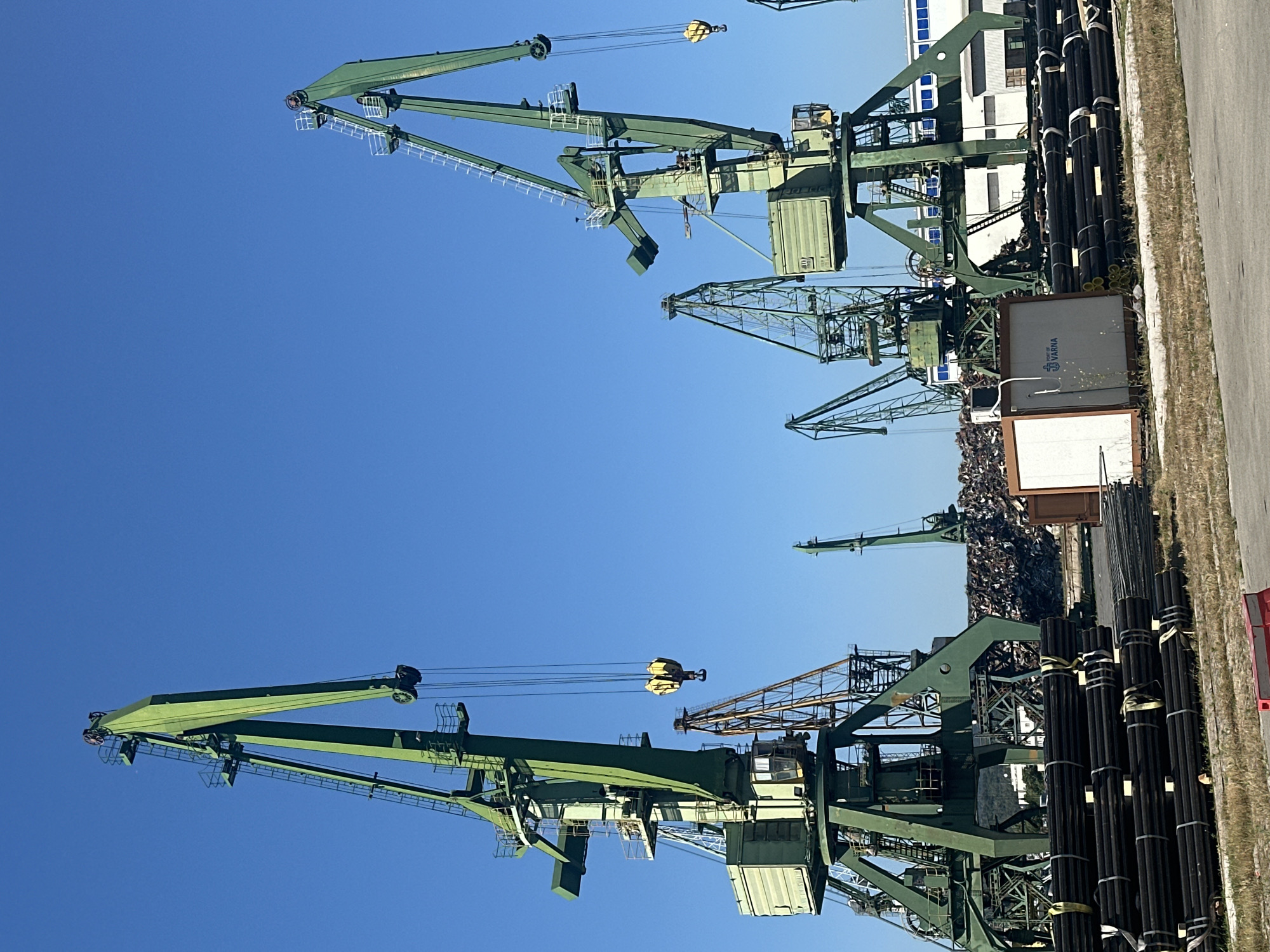 The image depicts an outdoor industrial scene, likely a port or shipyard, under a clear blue sky. In the foreground, neatly stacked bundles of dark, cylindrical pipes are visible, secured with straps. To the left and right, two large green harbor cranes dominate the frame. These cranes have extensive metallic structures, access ladders, and operator cabins. A yellow lifting hook hangs from the boom of the crane on the left. In the mid-ground, various other cranes, some appearing older and rust-colored, are present, along with a large pile of what appears to be scrap metal or debris. A white modular building with "PORT OF VARNA" branding is situated near the center. In the background, a long white industrial building with blue accents is visible. The ground is paved in some areas and covered with dry grass in others. There are no people visible in the image.
