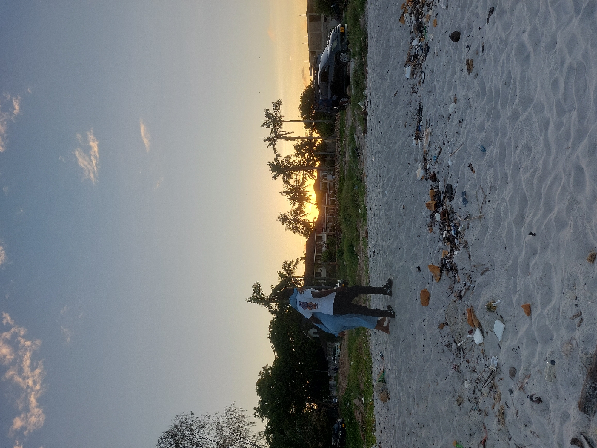 A landscape-oriented photo shows a beach scene near Dar es-Salaam, Tanzania, at sunset. Two people are standing on the sand, backlit by the setting sun. The beach has significant litter and debris along the edge. Palm trees line the shoreline with building silhouettes visible behind them. The sky is a gradient from blue to orange, with sparse clouds. A car is parked near the beach. The image is angled slightly to the right.