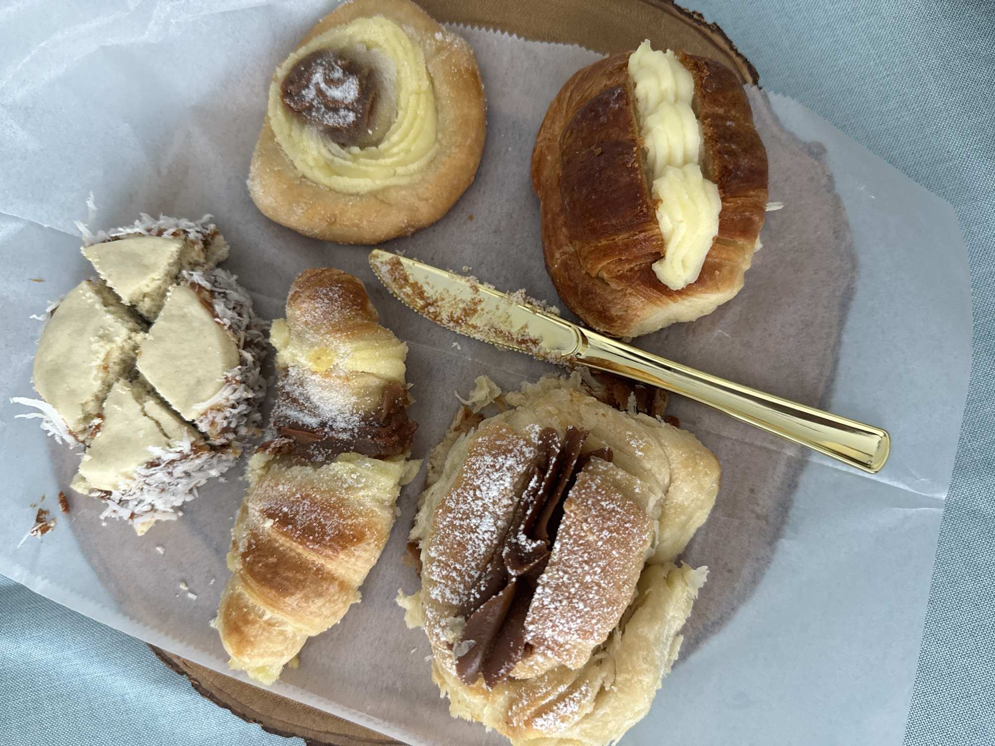 The image shows a variety of pastries arranged on a sheet of parchment paper, which sits atop a wooden surface. The pastries include an Alfajor cut into pieces and covered in shredded coconut, a filled spiral pastry, a croissant with cream filling, a small chocolate-filled croissant, and a larger croissant filled with chocolate and dusted with powdered sugar. A gold-colored butter knife rests among the pastries. The background includes a section of light blue fabric. The setting appears to be a bakery or food preparation area. There is no evidence in the image to suggest a location in Davie, Florida.