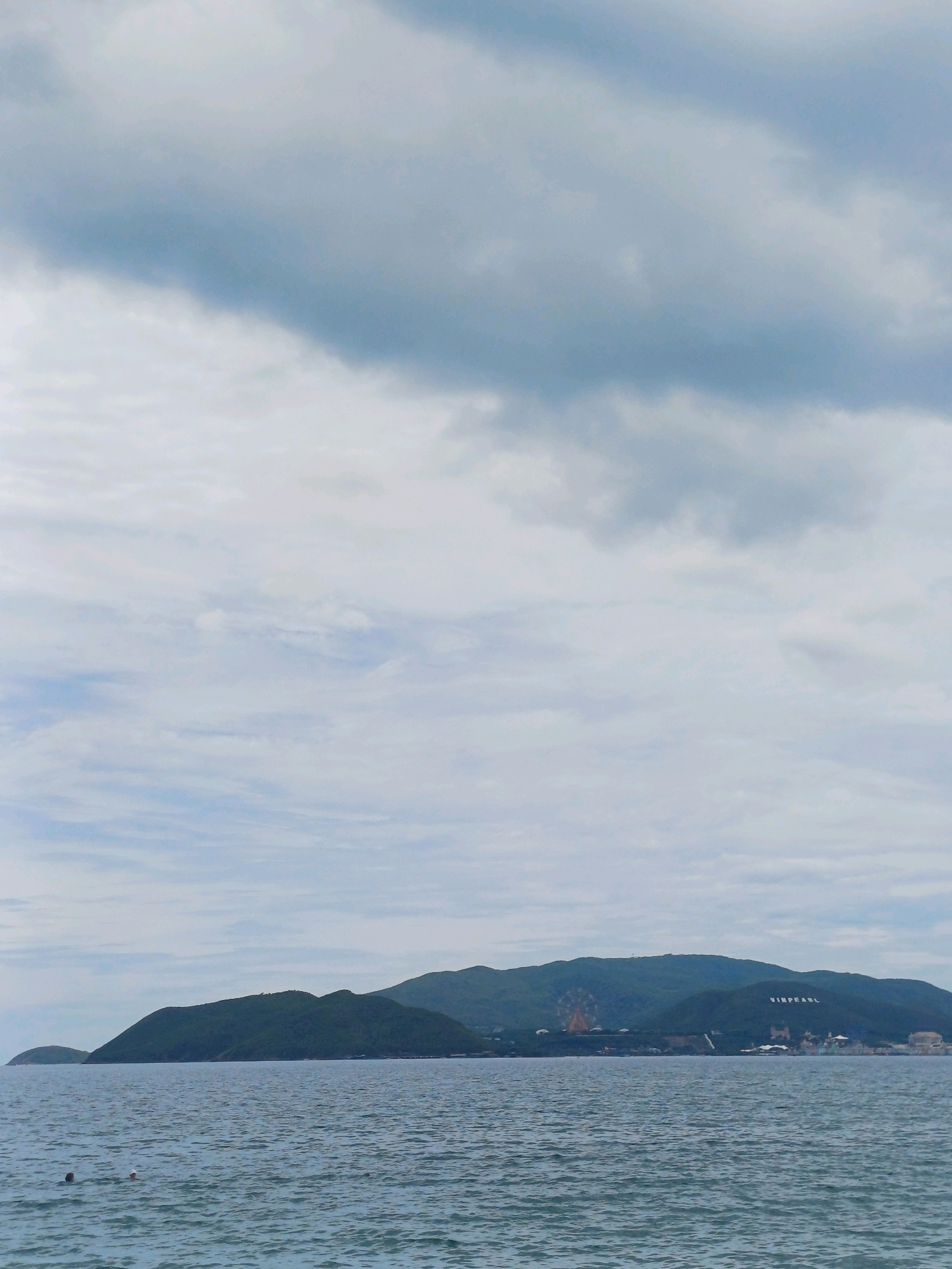 The image depicts a coastal landscape with a body of water in the foreground and a mountainous island in the background. The water is calm with slight ripples, and two people are visible swimming. The island is covered in lush green vegetation, with buildings and a ferris wheel visible near the summit. The word "VINPEARL" is written in large white letters on the side of the mountain. The sky is overcast with heavy clouds. The scene is consistent with views of Hon Tre Island in Nha Trang, Vietnam, home to the Vinpearl resort complex.