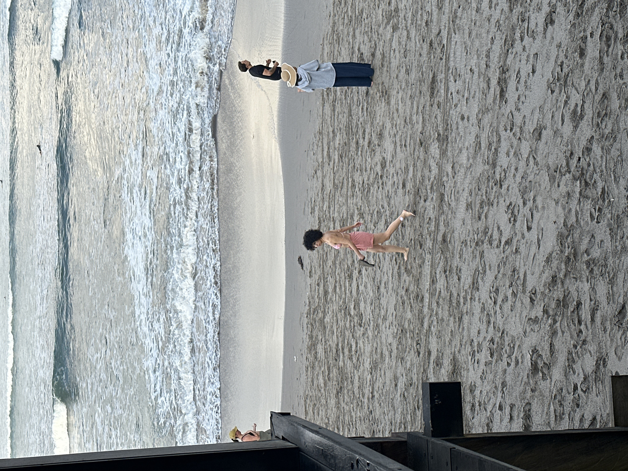 A wide shot captures a dusky beach in North Kuta, Indonesia, as waves gently break on the shore. In the foreground, a dark wooden structure partially obstructs the view on the left. On the sand, a woman in a pink outfit walks towards the right, holding a dark object in her hand. Further in the distance, another woman stands facing the ocean, wearing a wide-brimmed hat and a grey hooded jacket over dark pants. A man in a black t-shirt walks away from the camera, looking towards the right. The sand is textured with footprints, and a surfer is visible in the water.