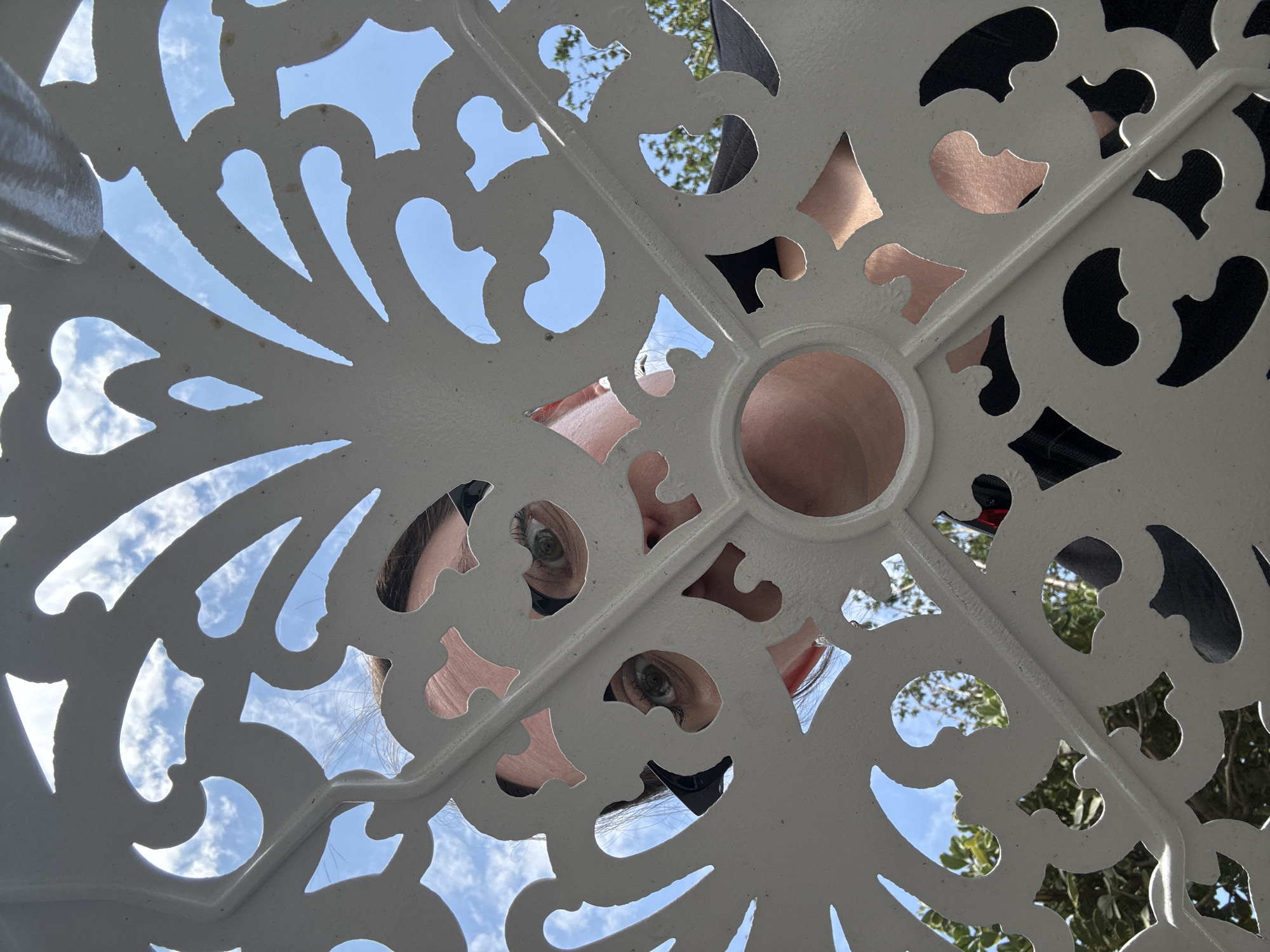 The image presents a low-angle perspective of a white, intricately designed metal table or structure, viewed against a backdrop of a blue sky with scattered clouds. A person's face is visible through the openings in the metalwork, primarily the eyes, nose, and parts of the forehead. The person seems to be looking directly at the camera. Sections of black clothing and hair are also visible. The environment suggests an outdoor setting with possible vegetation visible through the openings. The location in Moscow, Russia cannot be determined from the image alone.