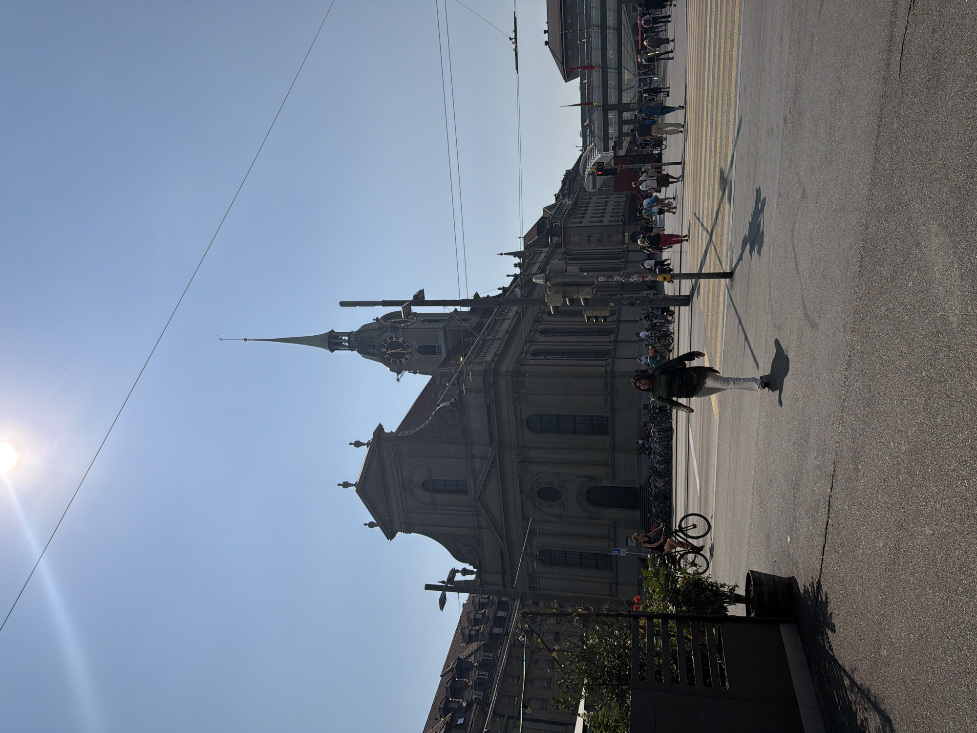 In Bern, Switzerland, the Zytglogge clock tower is visible in the background of this street scene. The tower is part of a historic building, possibly a church or cathedral, with a prominent spire and clock face. In the foreground, a woman walks across a crosswalk, while other pedestrians and cyclists navigate the city square. The sun is shining brightly, casting shadows on the pavement. The architecture suggests a European urban setting, with traditional buildings and a clear blue sky.