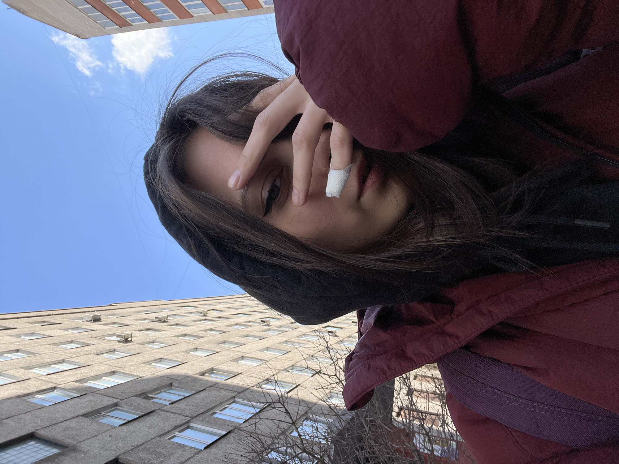 A low-angle, close-up shot shows a person with long brown hair, wearing a dark hooded sweatshirt and a burgundy winter coat, partially obscuring their face with their hand. A white bandage covers a finger. The person appears to be looking directly at the camera. The background consists of a multi-story concrete building with many windows and a clear blue sky with a few clouds visible in the upper left corner. This is not a photograph of Krasnoyarsk, Russia. The buildings are a different design to those in that Russian city.